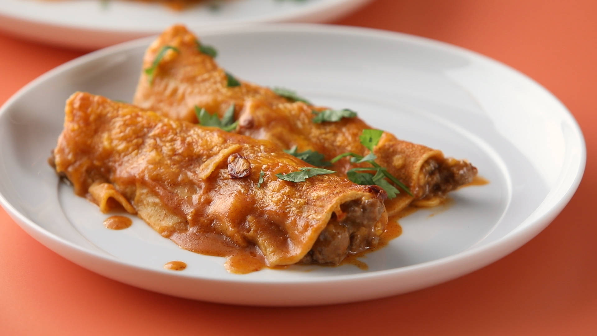 兩條 enchiladas 撒香菜 壁紙 — 兩個捲好的墨西哥捲餅，淋上濃郁的紅醬和新鮮香菜，放在白色盤子上，背景是溫暖的橙色。