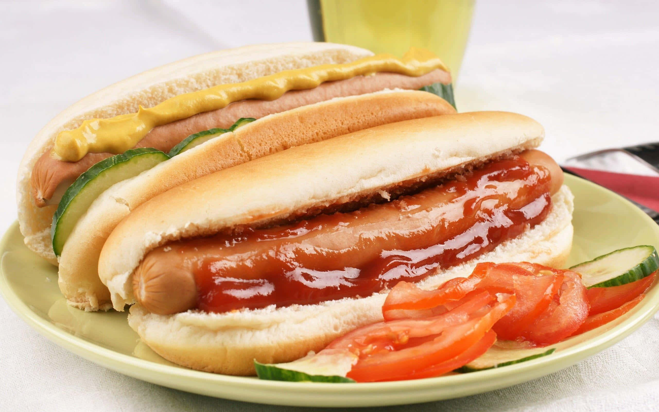 Hot Dog 桌布 壁紙 — 兩條多汁的熱狗，上面淋上茄汁、芥末和新鮮蔬菜，放在綠色碟上，背景有一杯飲品。