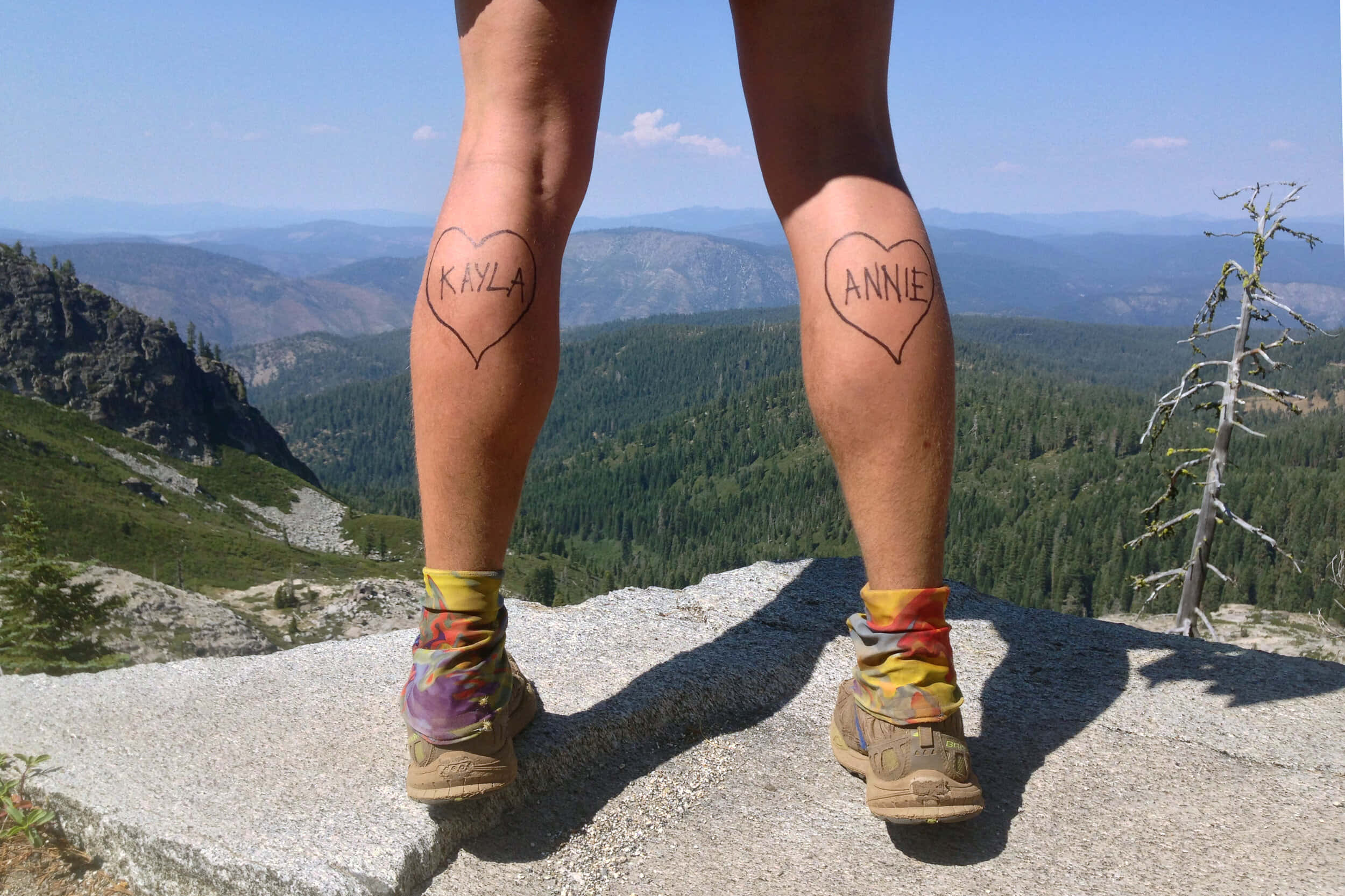 Hiking 桌布 壁紙 — 兩條帶有心形紋身、寫著 Kayla 和 Annie 的腿，站在岩石山頂上，俯瞰著晴朗藍天下廣闊的森林山谷。