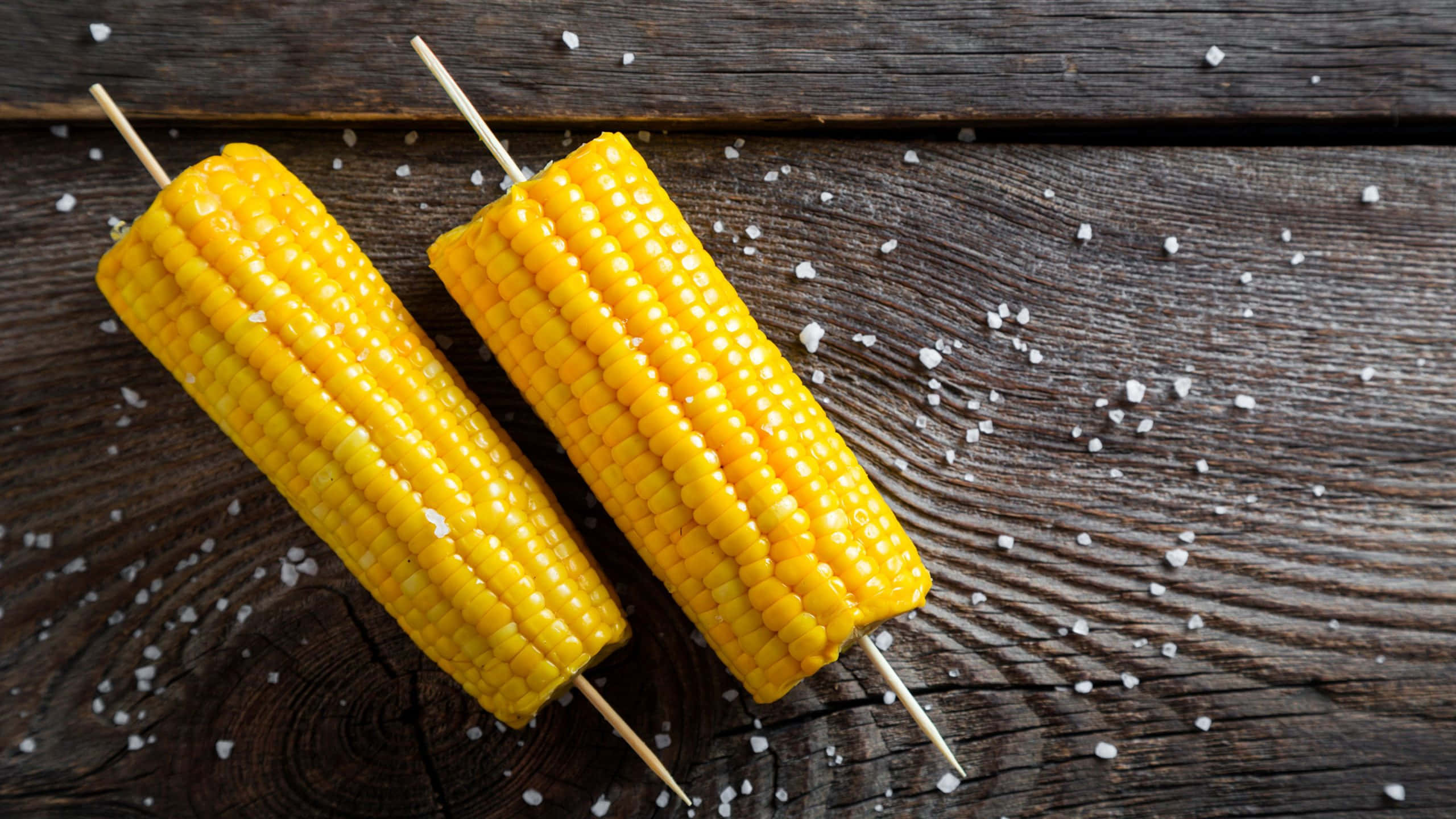 Corn 桌布 壁紙 — 兩條鹽水粟米串放在經風化的木質表面上，周圍散落著粗鹽。
