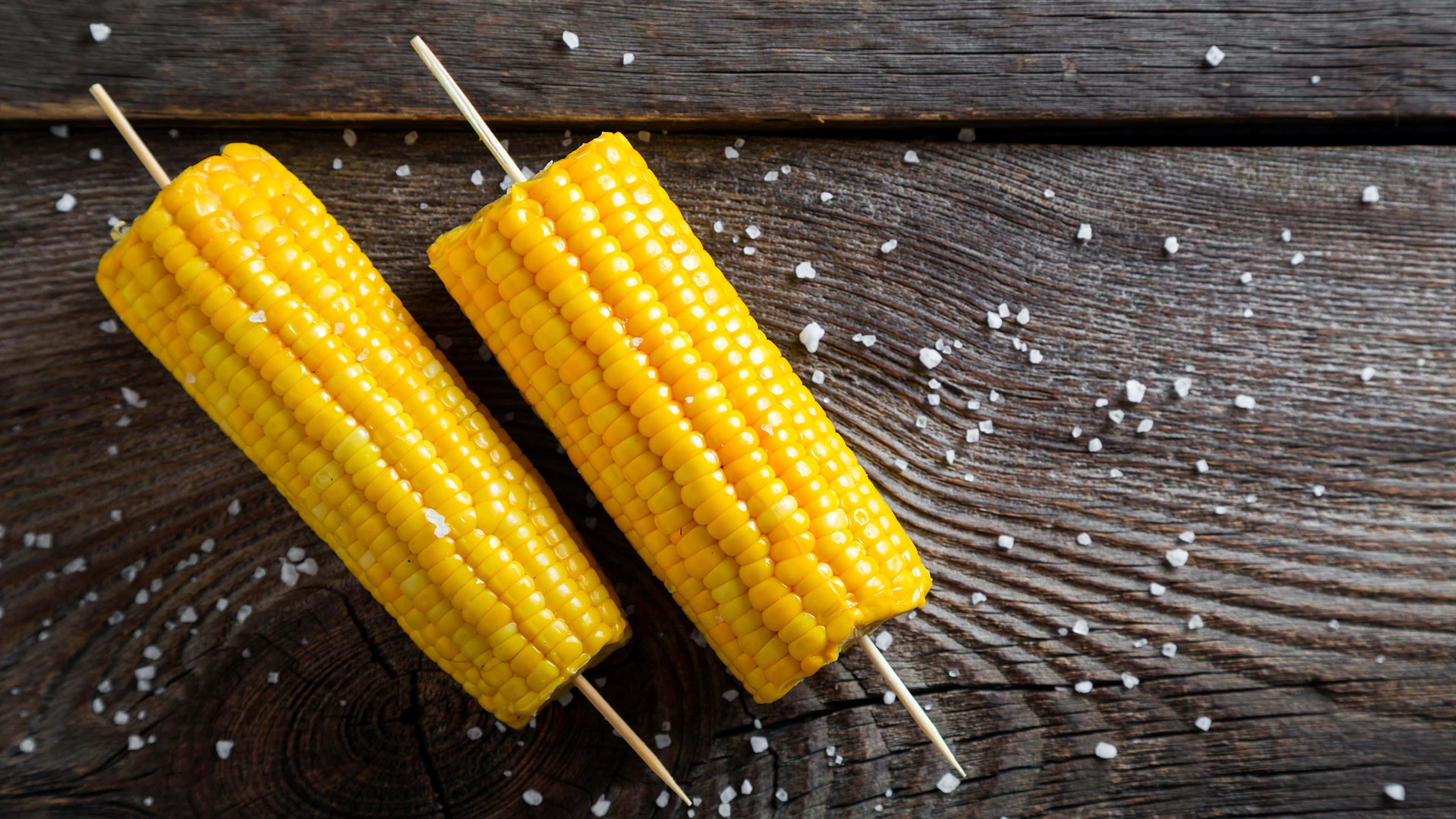 Yellow Corn 桌布 壁紙 — 兩條鹽烤粟米串放在經風化的木質表面上，周圍散落著粗鹽。