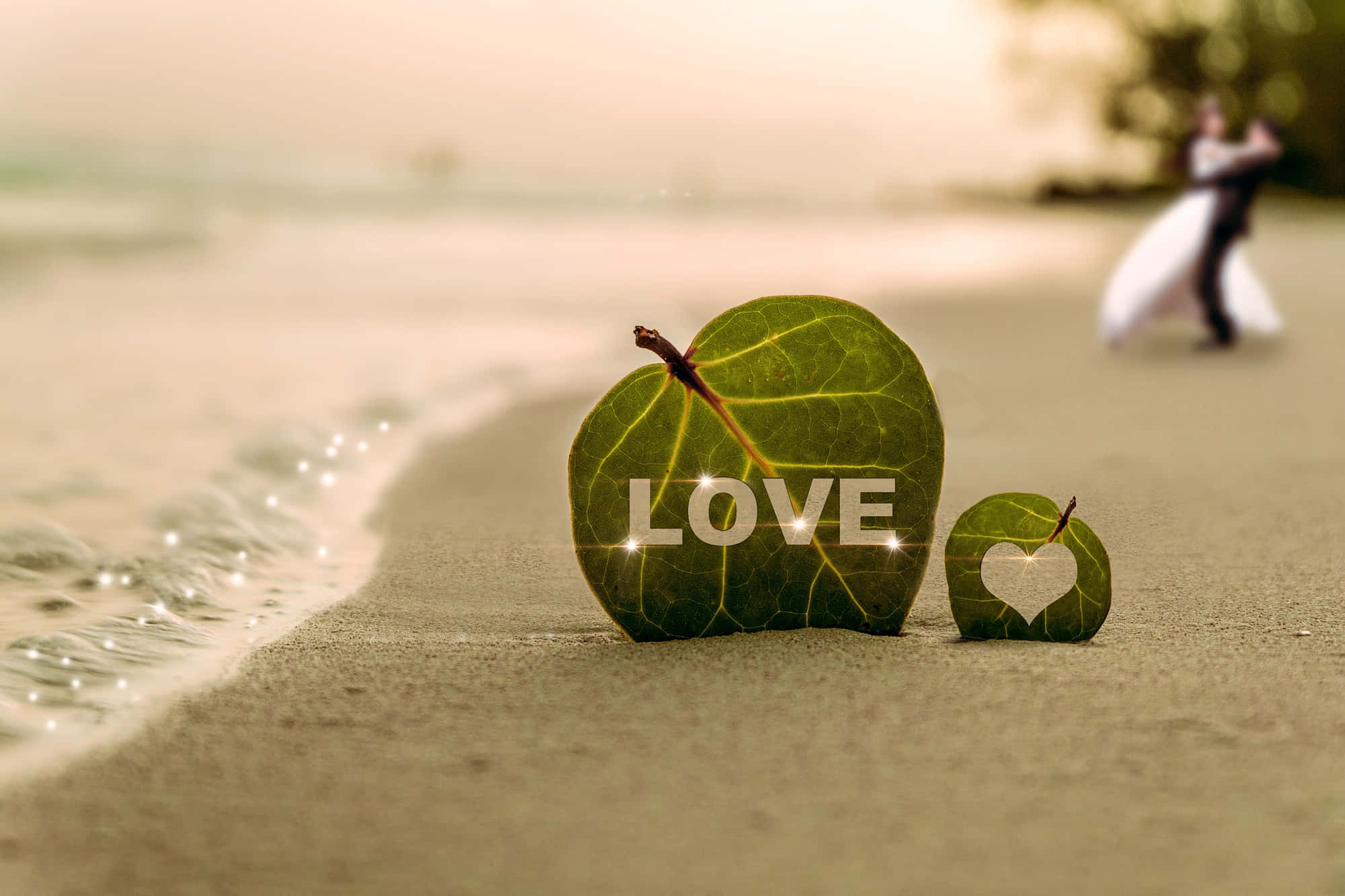Beach Love 桌布 壁紙 — 兩片心形葉片帶有「LOVE」文字在沙灘上發光，背景中一對模糊的情侶在黃金時刻跳舞。