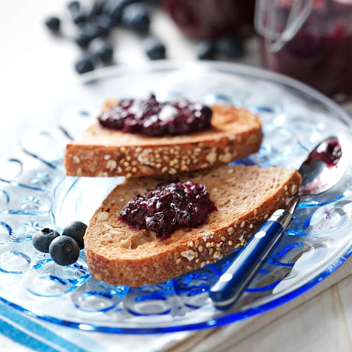Blueberry Jam 桌布 壁紙 — 兩片籽粒全麥多士，上面塗有光澤的藍莓果醬，放在華麗的藍色玻璃盤上，配以新鮮漿果和勺子。