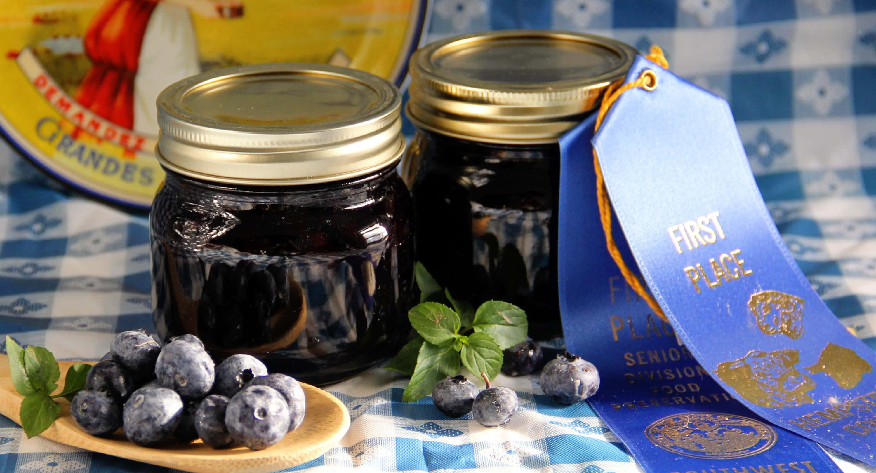 Blueberry Jam 桌布 壁紙 — 兩瓶深色藍莓醬玻璃罐放在藍色格紋布上，旁邊有新鮮莓果和薄荷，藍色絲帶宣告第一名。