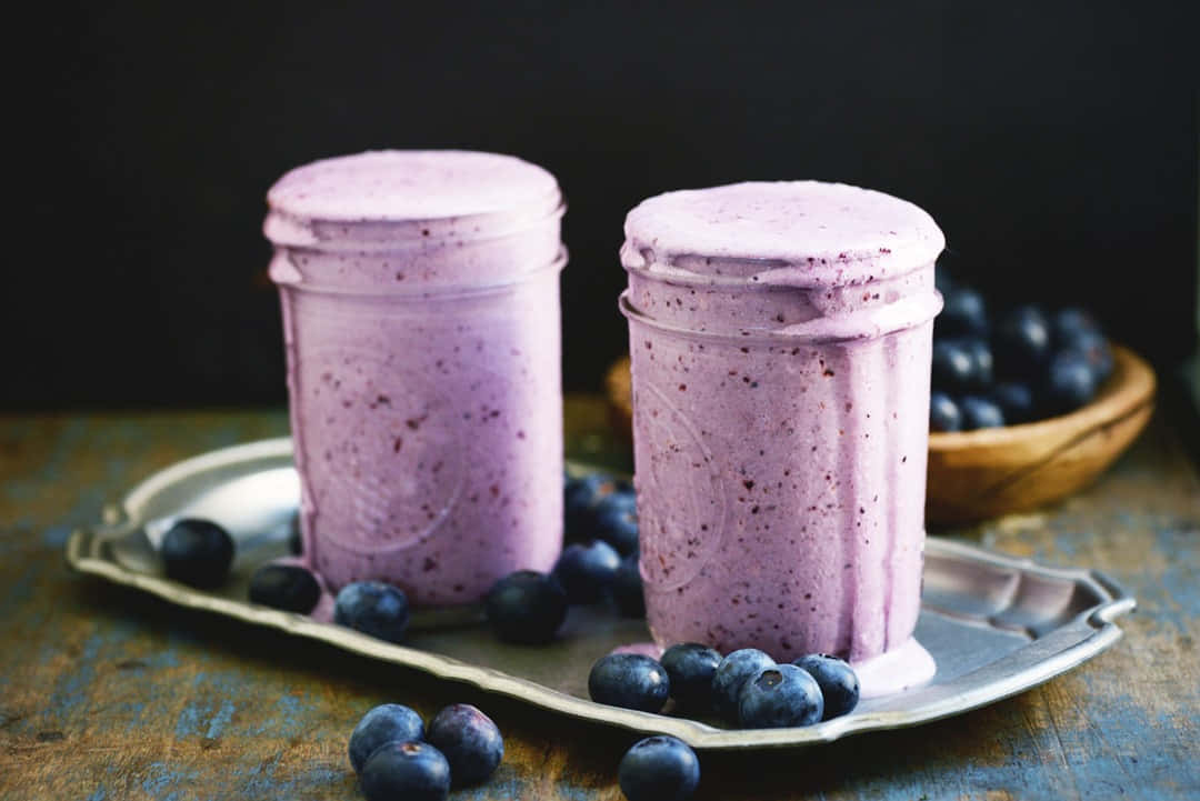 Blueberry Smoothie 桌布 壁紙 — 兩罐充滿活力的紫色藍莓冰沙放在銀托盤上，周圍是新鮮藍莓，背景是深色鄉村風格。