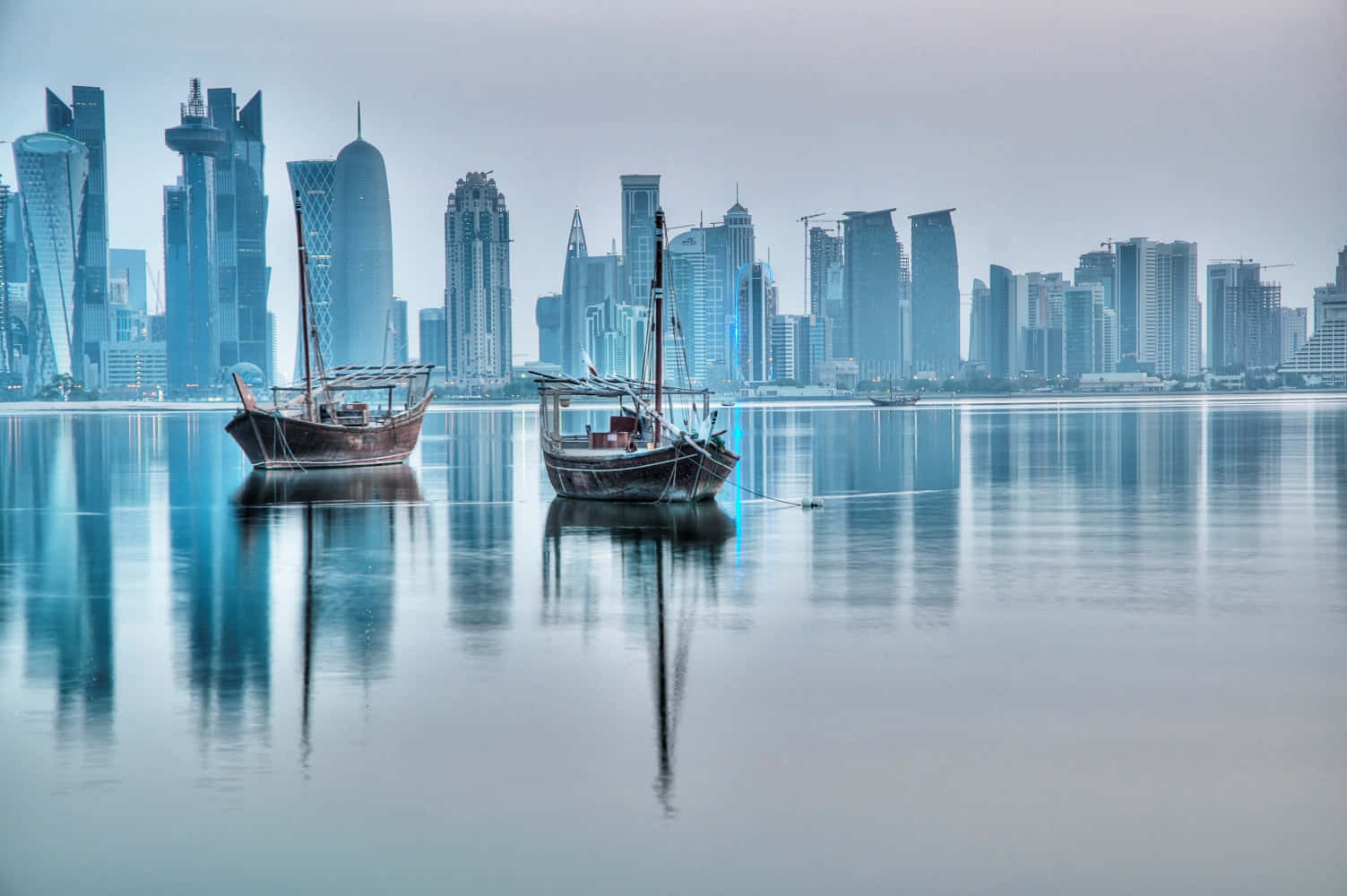 Qatar 桌布 壁紙 — 兩艘傳統木製單桅帆船在平靜的水面上悠然漂浮，水面倒映著霧氣籠罩的天空下未來主義的城市天際線。