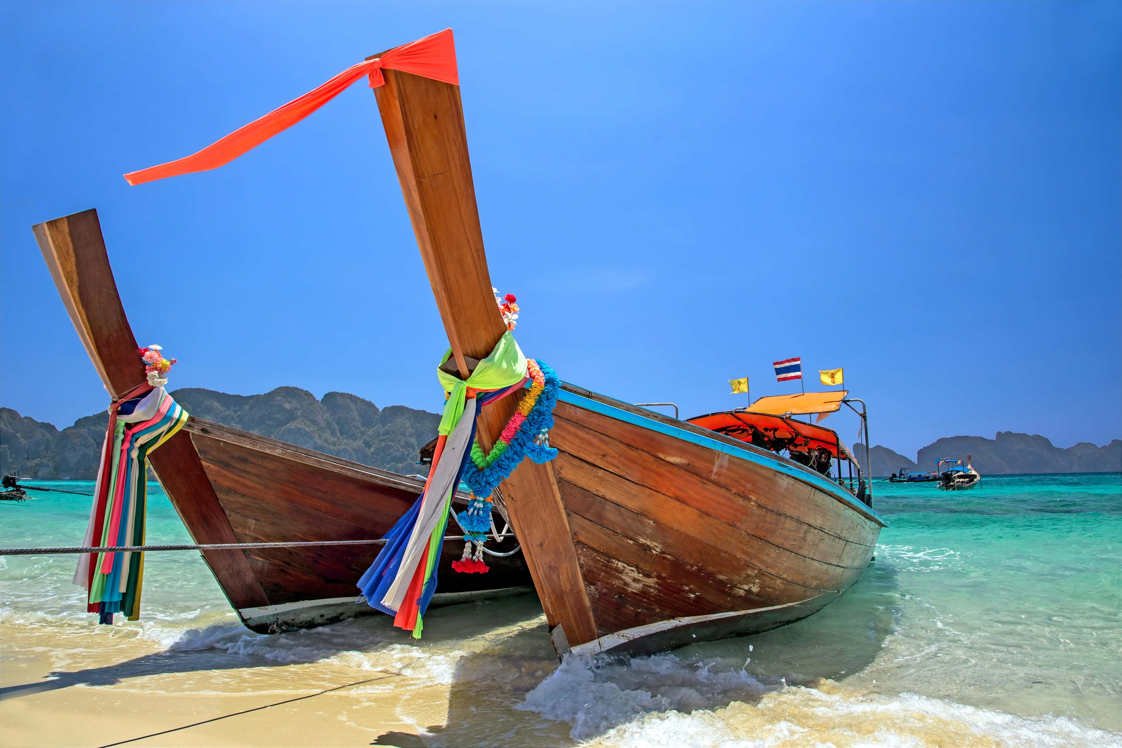 Boating 桌布 壁紙 — 兩艘裝飾著彩色絲帶的傳統泰國長尾船，在清澈藍天下以崎嶇山脈為背景，停泊在淺藍綠色的海水中。