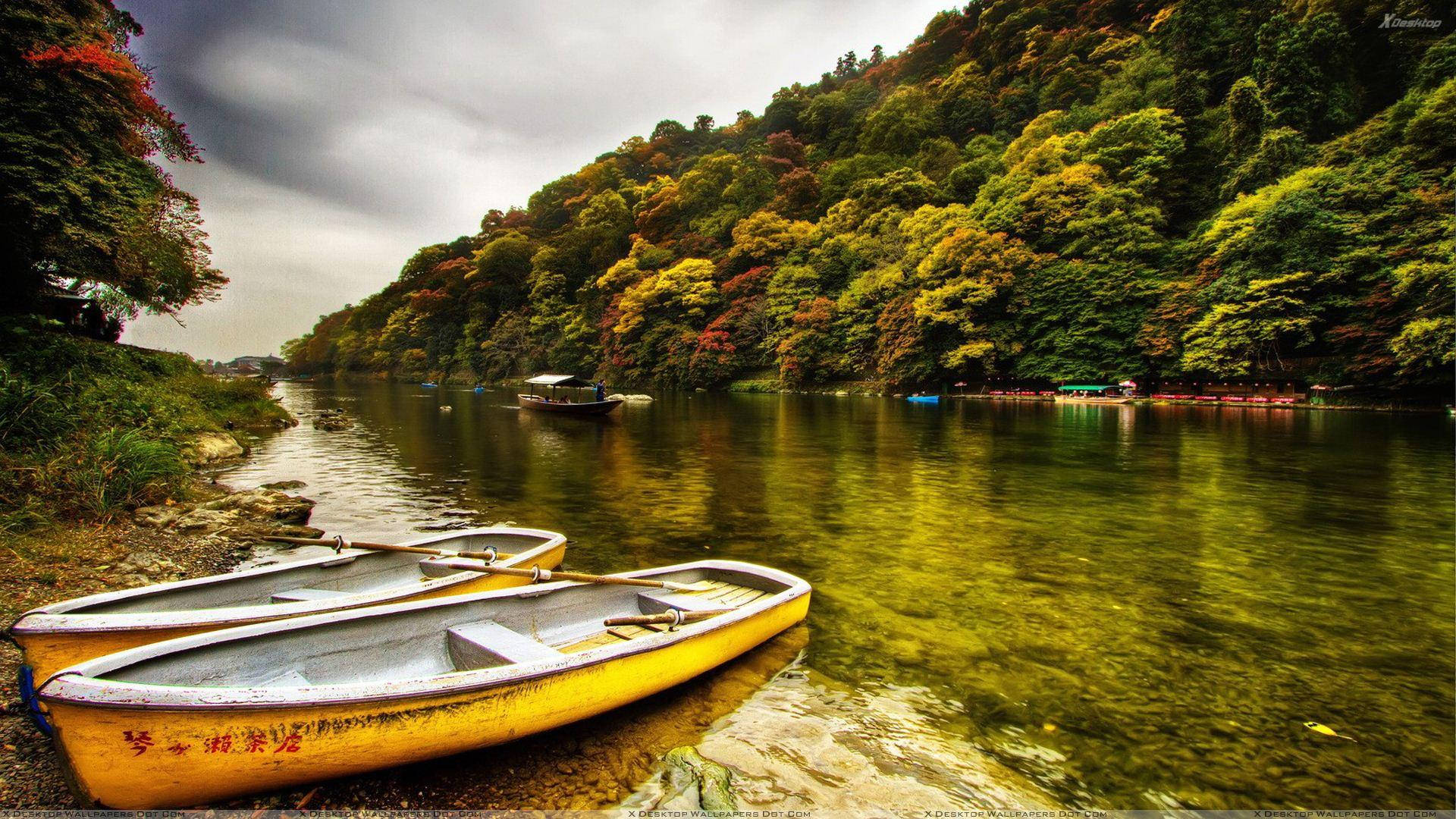 river 桌布 壁紙 — 兩艘風化的黃色划艇停泊在長滿苔蘚的河岸上，上方是戲劇性的多雲天空，周圍環繞著充滿活力的秋葉。