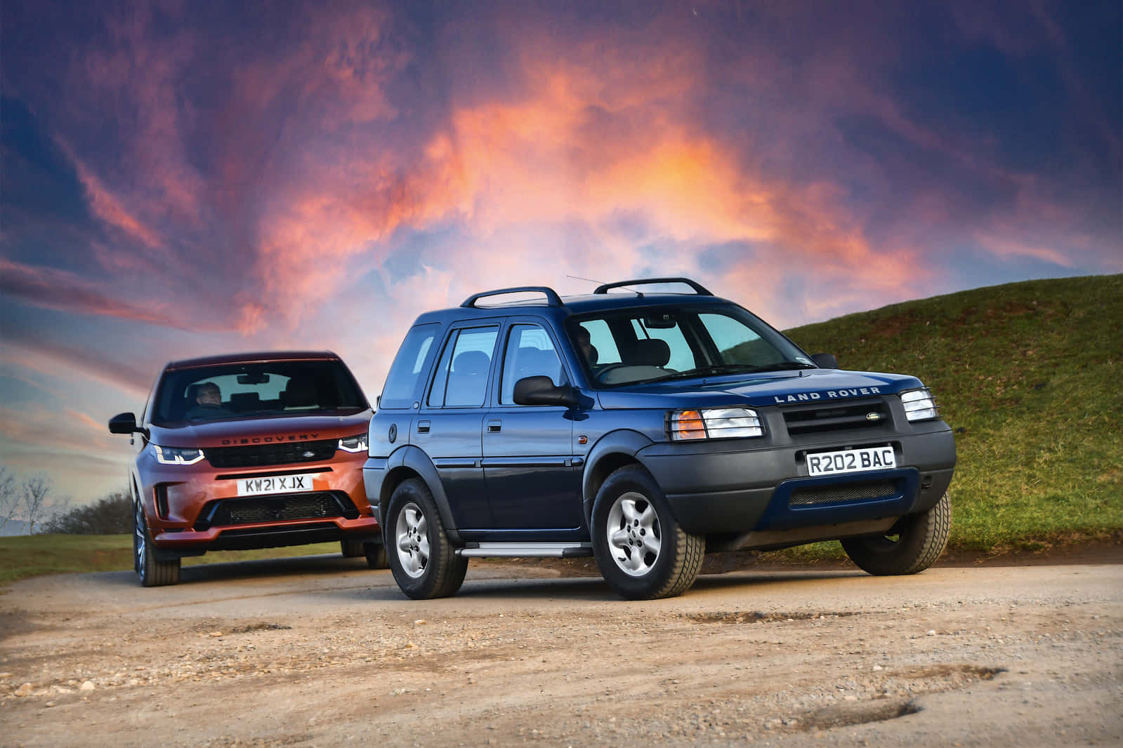 Land Rover Freelander 桌布 壁紙 — 兩輛 Land Rover Discovery SUV 停泊在一條泥路上，背景是壯麗的日落天空，帶有火橙色和紫色的雲彩。