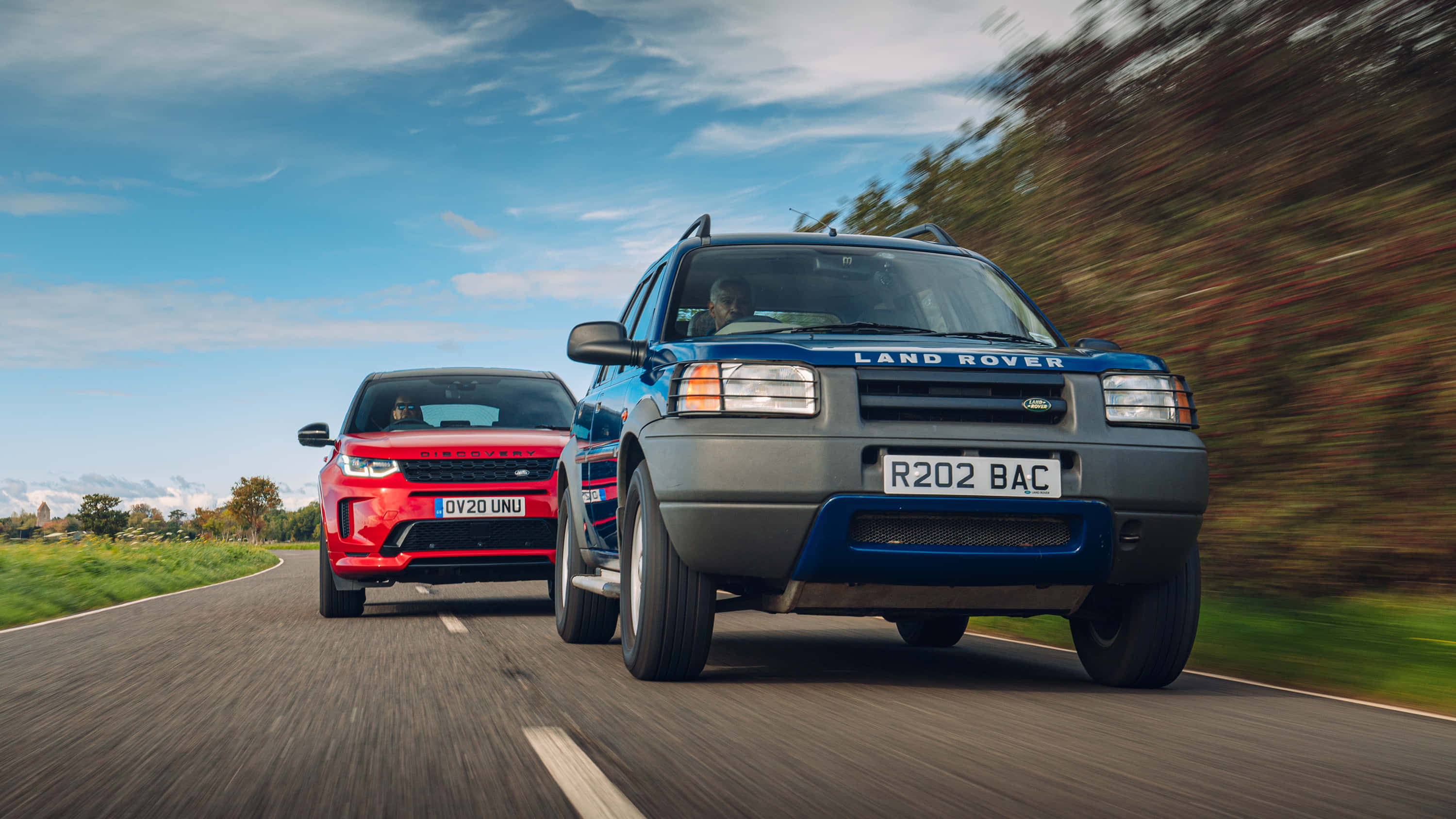 Land Rover Freelander 桌布 壁紙 — 兩輛Land Rover SUV在明亮的藍天下沿著蜿蜒的鄉村道路高速行駛，樹木呈現動態模糊效果。