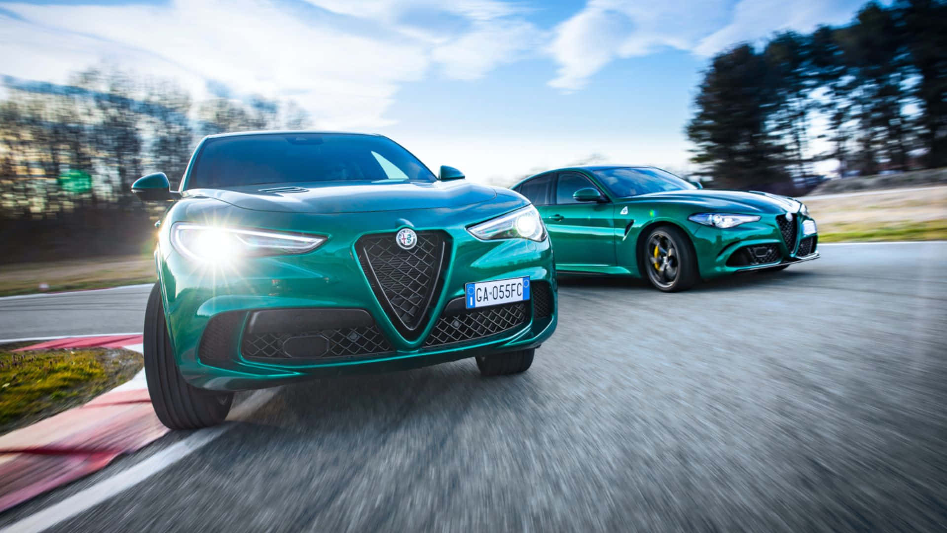 Alfa Romeo Stelvio 桌布 壁紙 — 兩輛充滿活力的綠色愛快羅密歐跑車在蜿蜒的道路上並排行駛，帶有模糊的動態和明亮的陽光。