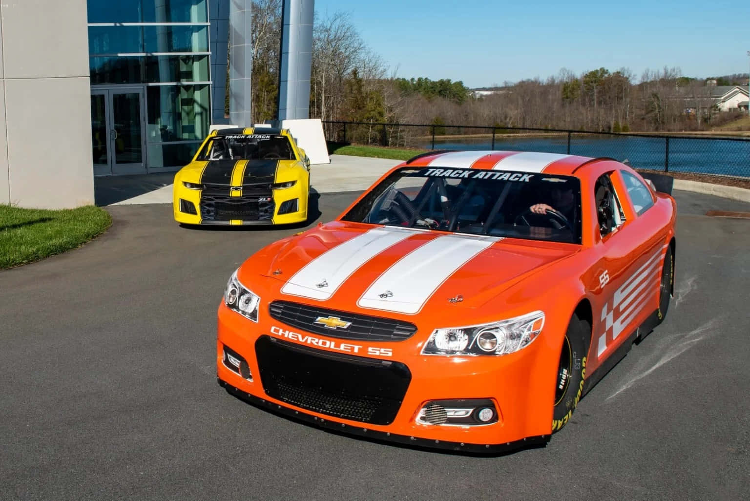 Car Racing 桌布 壁紙 — 兩輛充滿活力的賽車，橙色和黃色帶有白色條紋，在陽光明媚的日子停在一棟現代玻璃建築外。