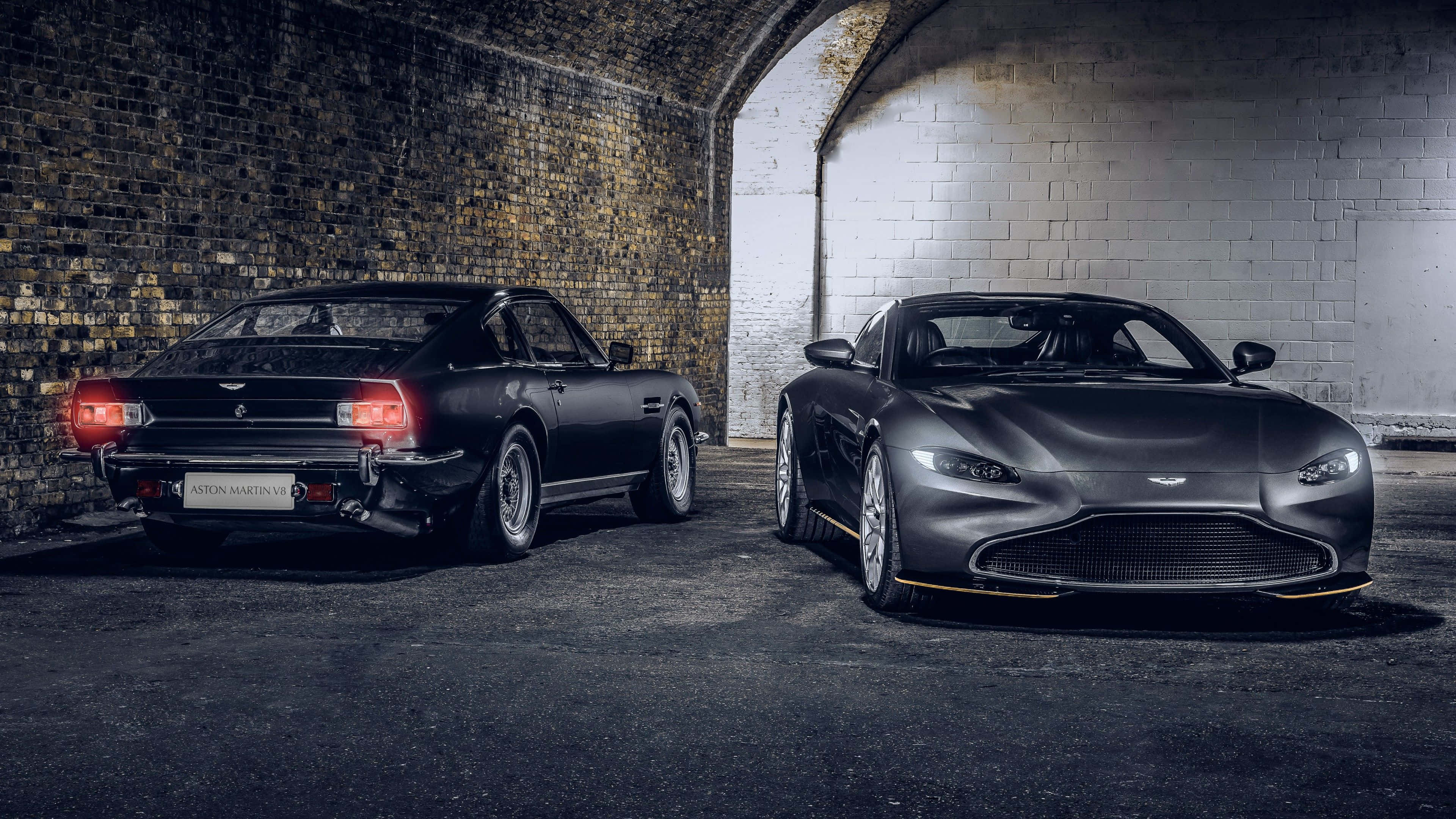 Aston Martin V12 Vantage 桌布 壁紙 — 兩輛啞光黑色的Aston Martin跑車停放在光線昏暗的磚砌隧道中，帶有戲劇性的燈光效果。