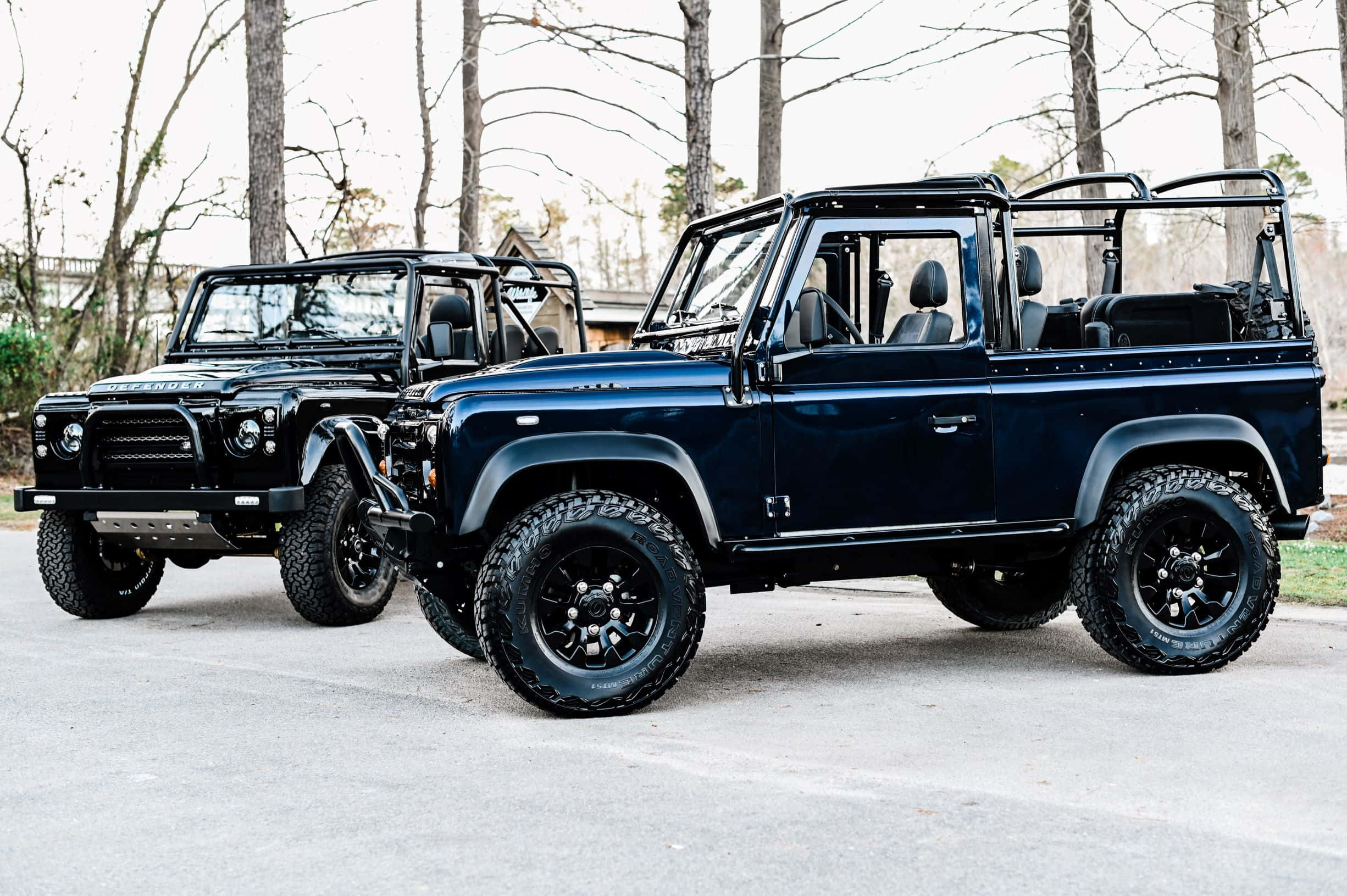 Land Rover Defender 桌布 壁紙 — 兩輛堅固的黑色 Land Rover Defender 並排停泊在鋪砌路面上，背景有光禿禿的樹木。