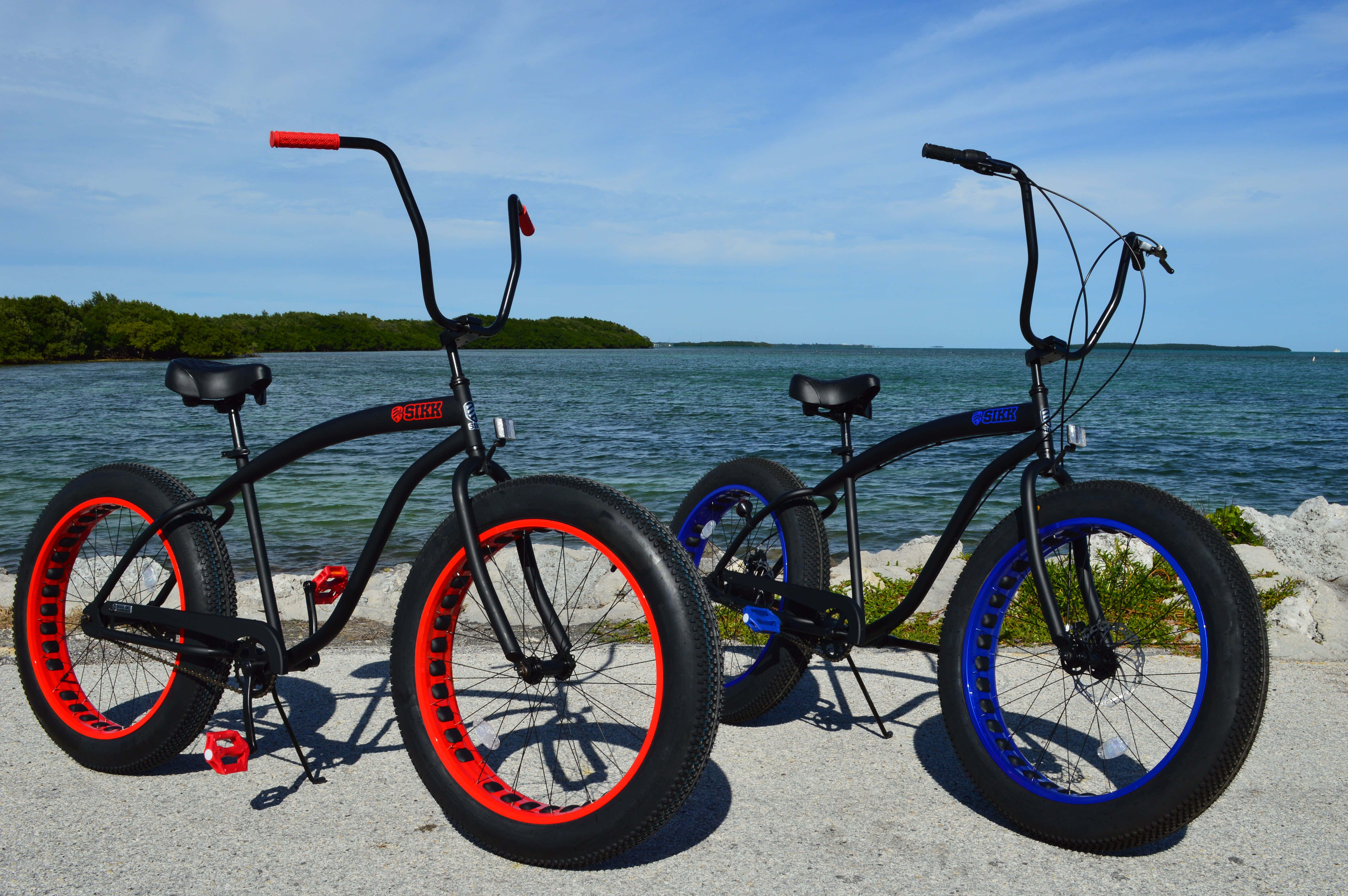 Beach Cruiser 桌布 壁紙 — 兩輛大膽的黑色胖胎車，帶有紅色和藍色輪圈，站在清澈天空下平靜藍水旁的礫石岸邊。