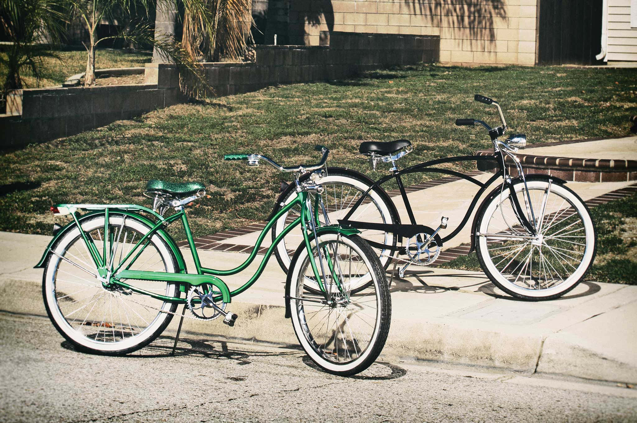 Beach Cruiser 桌布 壁紙 — 兩輛復古單車，一綠一黑，停放在陽光普照下草地旁的行人路上。