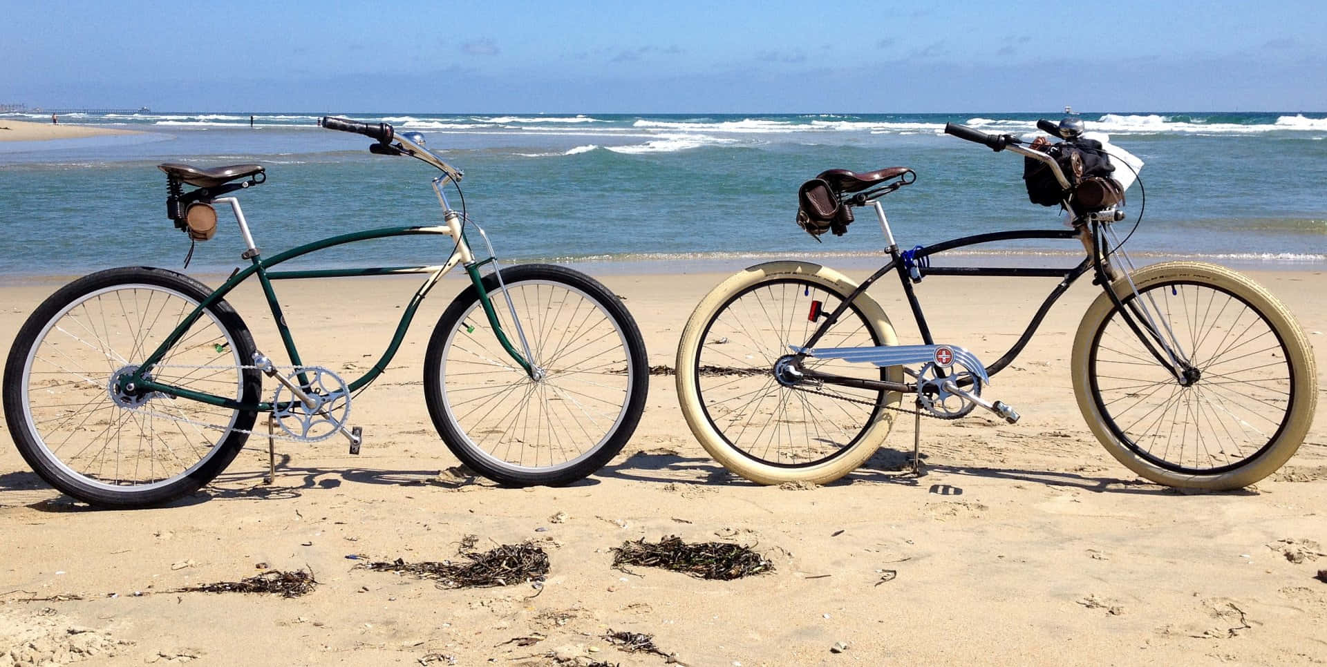 Beach Cruiser 桌布 壁紙 — 兩輛復古自行車，車架為奶油色和綠色，並排停放在陽光明媚的沙灘上，面向海洋的波浪。