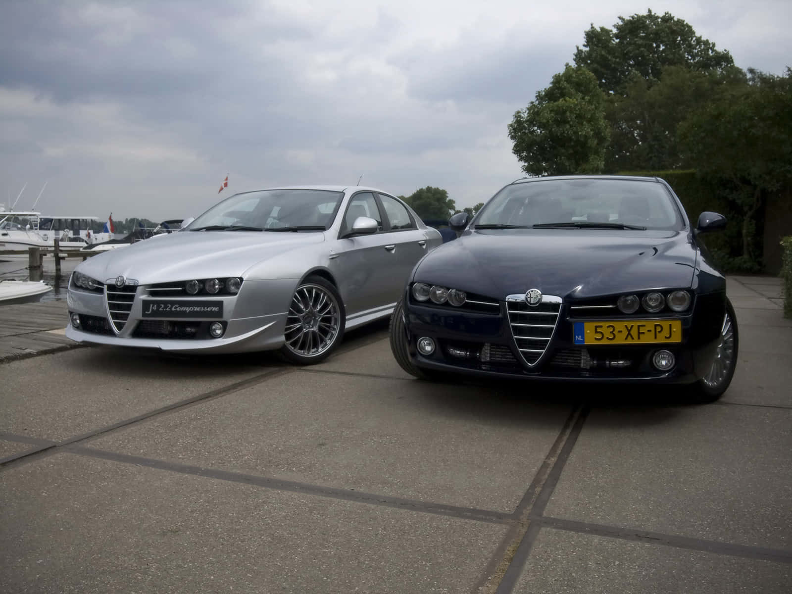 Alfa Romeo 159 桌布 壁紙 — 兩輛時尚的Alfa Romeo房車，銀色和黑色，並排停泊在鋪砌的碼頭上，背景陰天，有船隻和樹木。