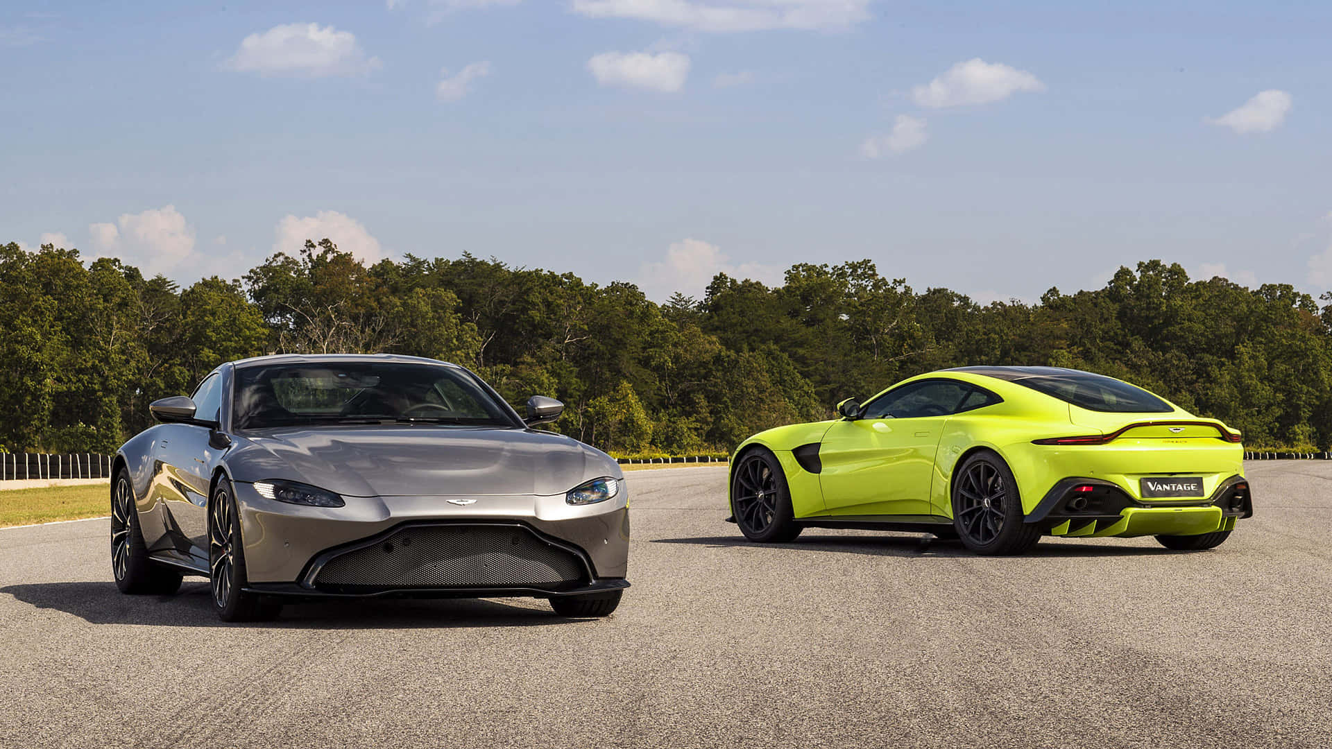 Aston Martin Vantage 桌布 壁紙 — 兩輛時尚的Aston Martin Vantage跑車，一輛銀色，一輛螢光黃色，停在瀝青賽道上，背景有樹木和藍天。