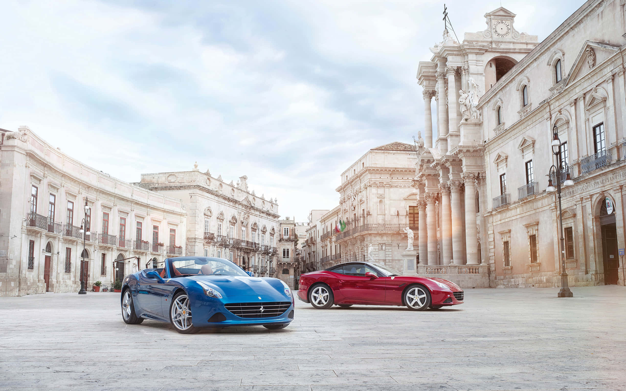 Ferrari California T 桌布 壁紙 — 兩輛時尚的Ferrari跑車藍色和紅色停放在一個宏偉的歐洲廣場上，周圍是歷史建築，天空柔和多雲。