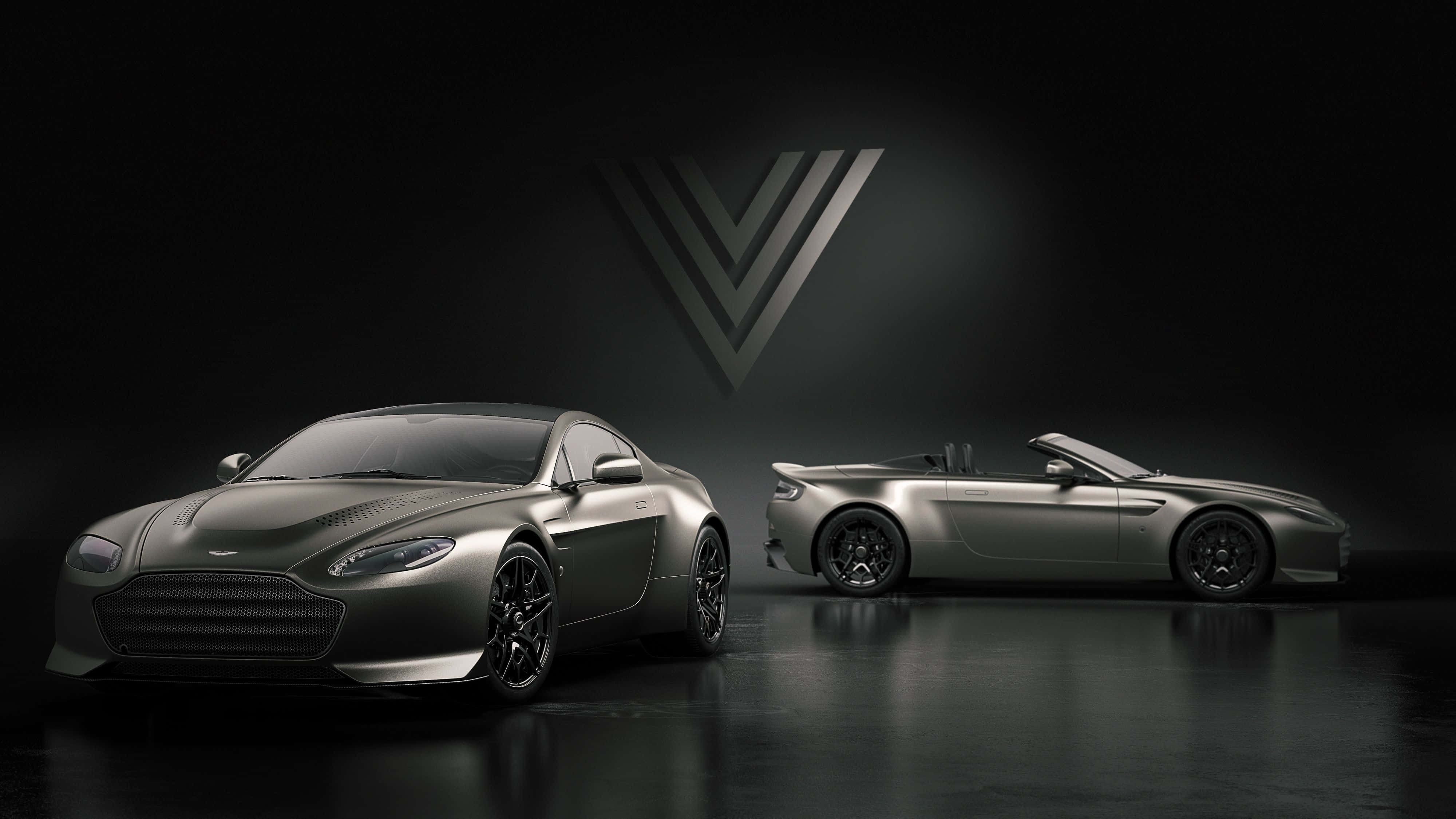 Aston Martin V12 Vantage 桌布 壁紙 — 兩輛時尚的啞光灰色 Aston Martin 跑車在黑暗的攝影棚背景下，於戲劇性的聚光燈下閃耀，並帶有發光的 V 形標誌。