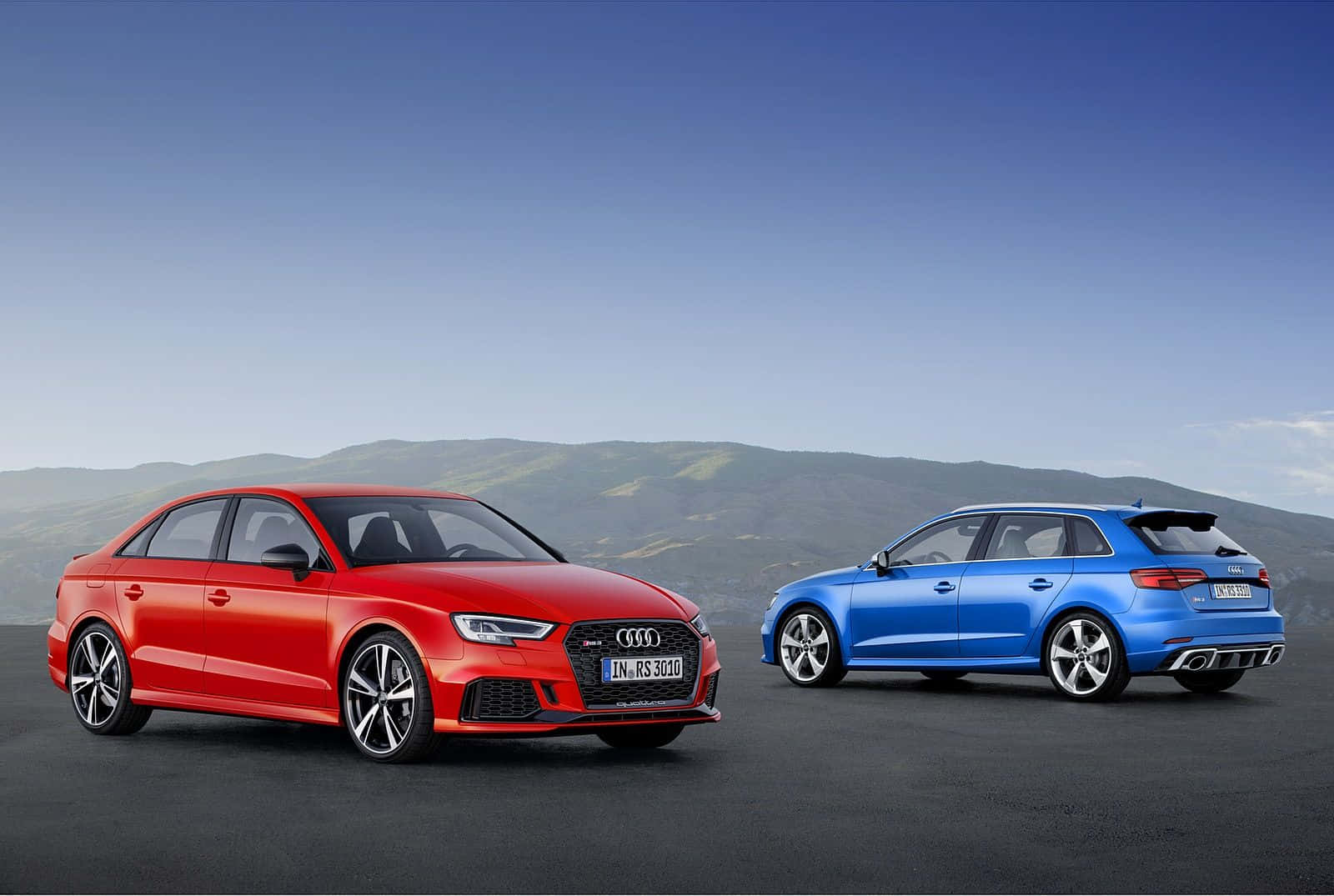 Audi Rs3 桌布 壁紙 — 兩輛時尚的奧迪RS3房車，一紅一藍，停在深色路面上，背景是連綿起伏的綠色山丘和晴朗的藍天。