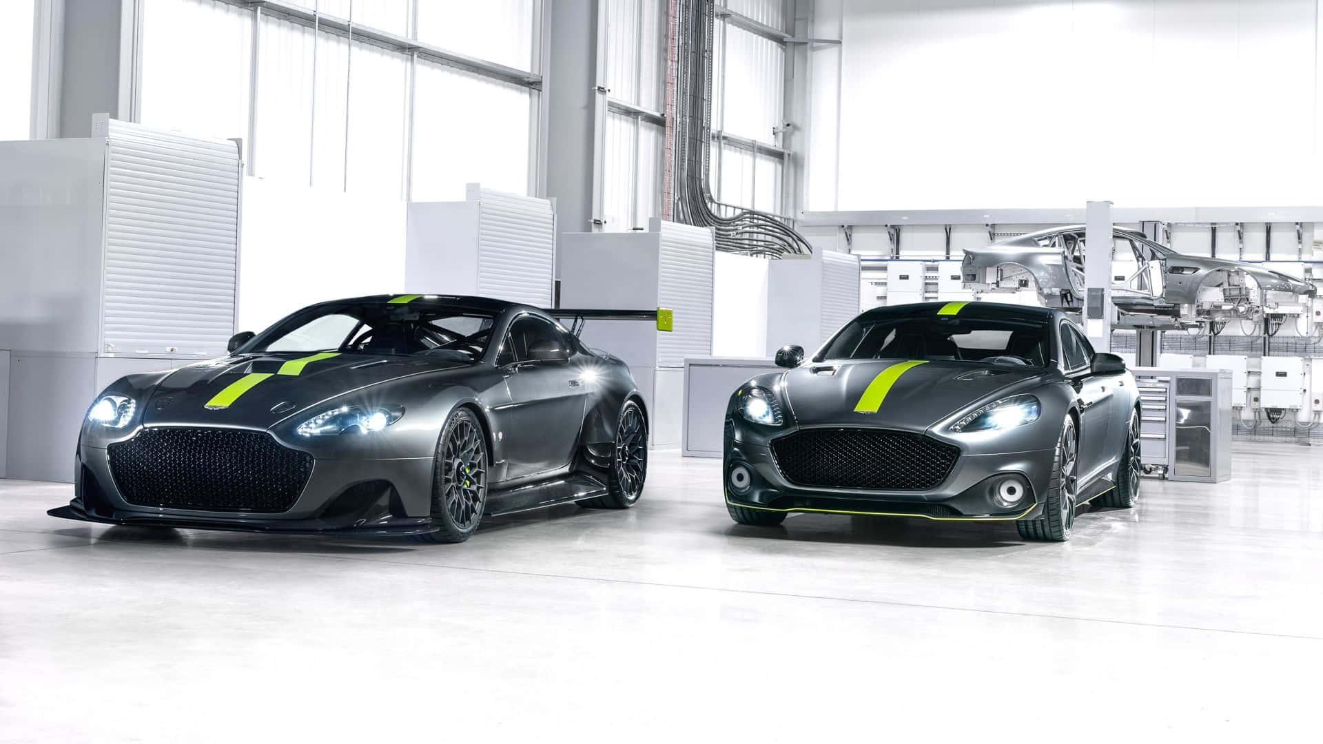 Aston Martin Rapide S 桌布 壁紙 — 兩輛時尚的灰色Aston Martin賽車帶有螢光綠色條紋，在簡約工業車庫的明亮燈光下閃閃發光。