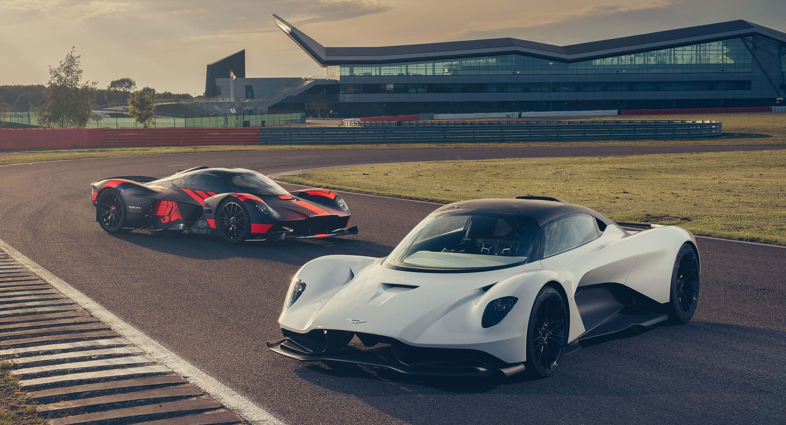 Aston Martin Valkyrie 桌布 壁紙 — 兩輛時尚超跑，一白一紅黑，停在賽道上，背景是現代建築。