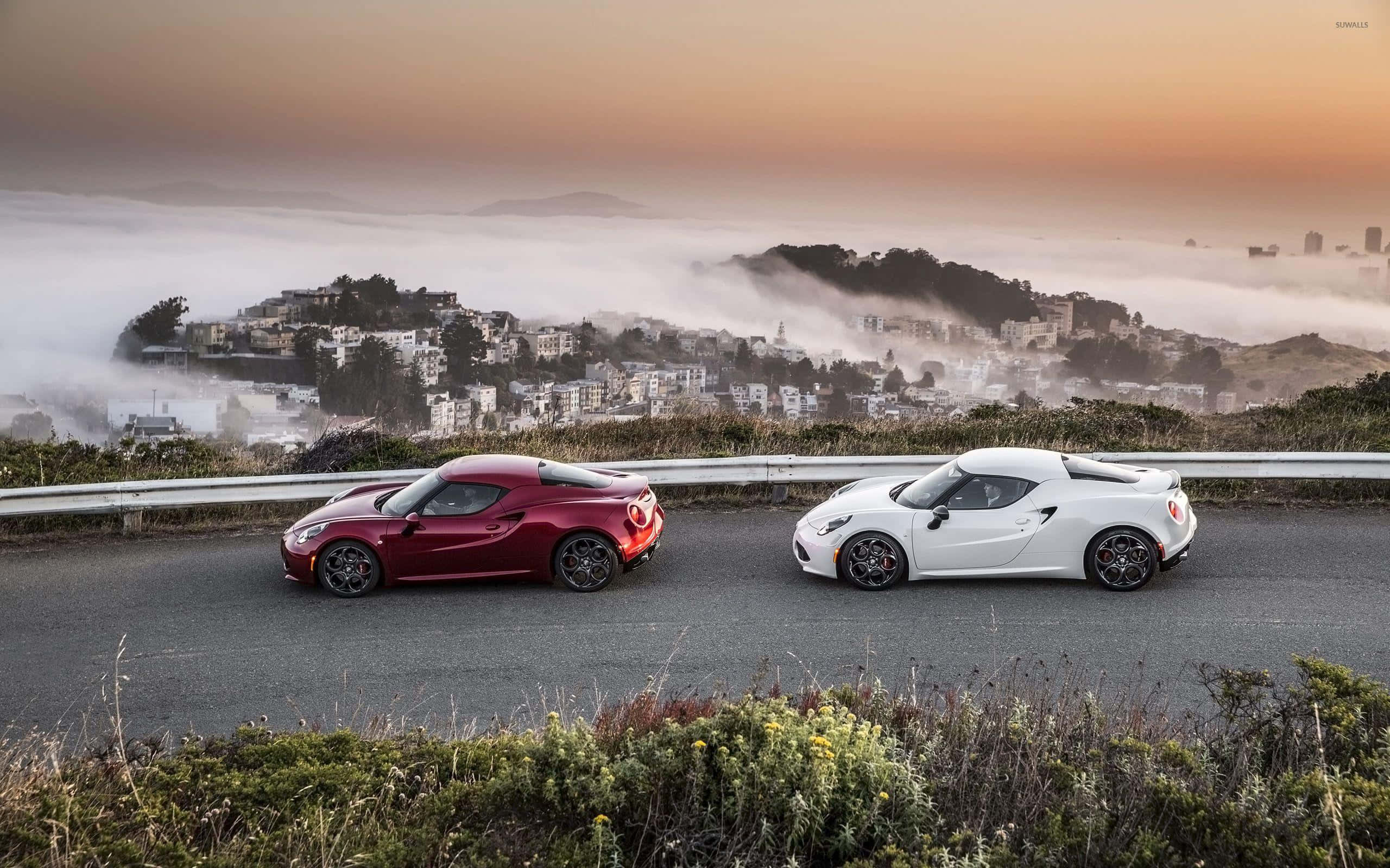 Alfa Romeo 4c 桌布 壁紙 — 兩輛時尚跑車，一輛紅色和一輛白色愛快羅密歐，並排停在沿海公路上，俯瞰日出時霧氣瀰漫的城市景觀。