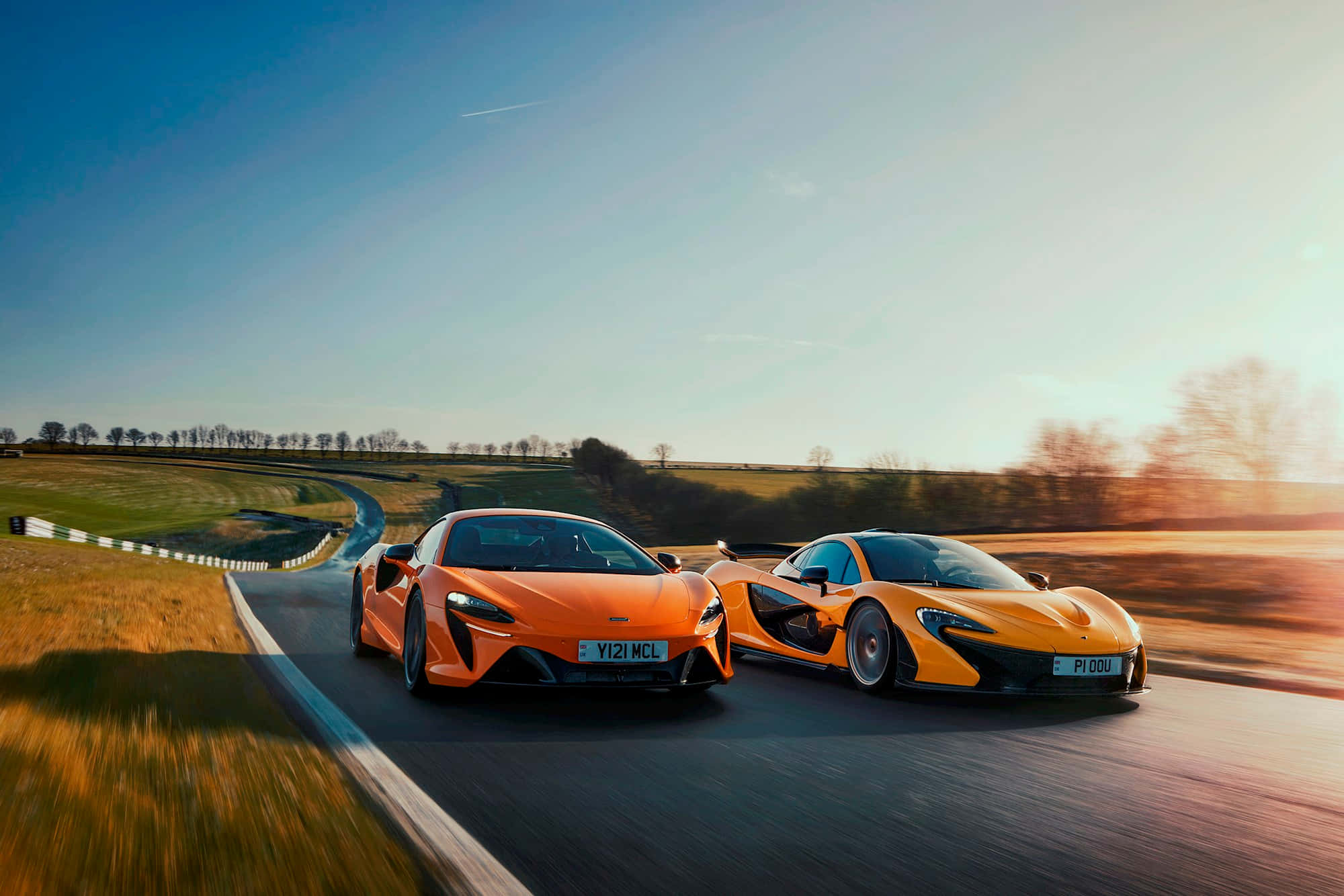 麥拿侖 桌布 壁紙 — 兩輛橙色McLaren超級跑車在黃金時段並排行駛在蜿蜒的鄉村道路上。
