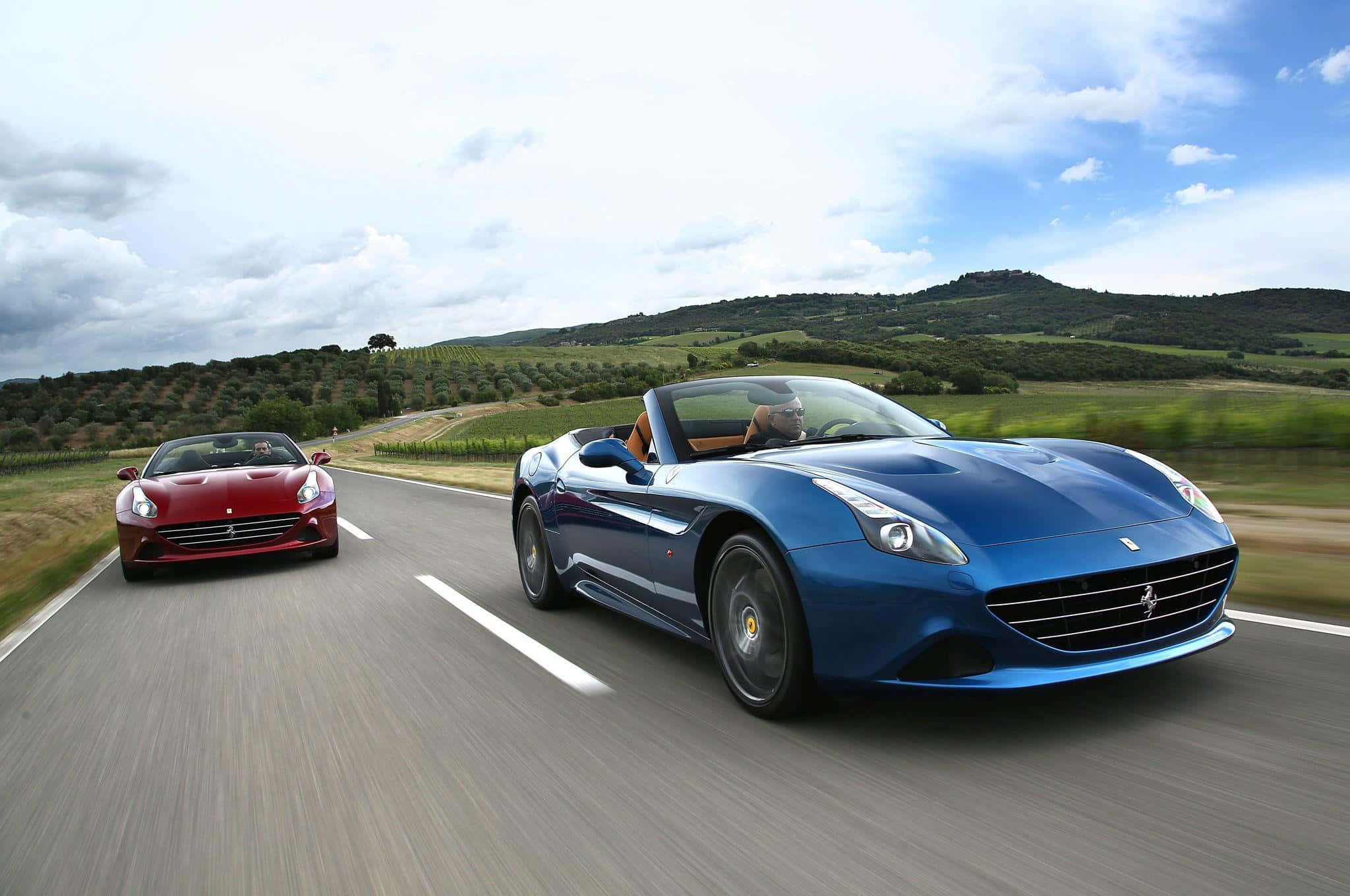 Ferrari California T 桌布 壁紙 — 兩輛法拉利開篷車在部分多雲的天空下沿著蜿蜒的鄉村道路競速。