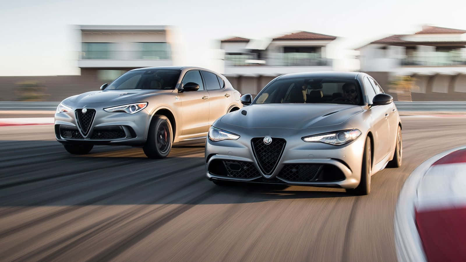 Alfa Romeo Stelvio 桌布 壁紙 — 兩輛流線型銀色愛快羅密歐並排馳騁在陽光普照的賽道上，背景有模糊的動態和現代房屋。