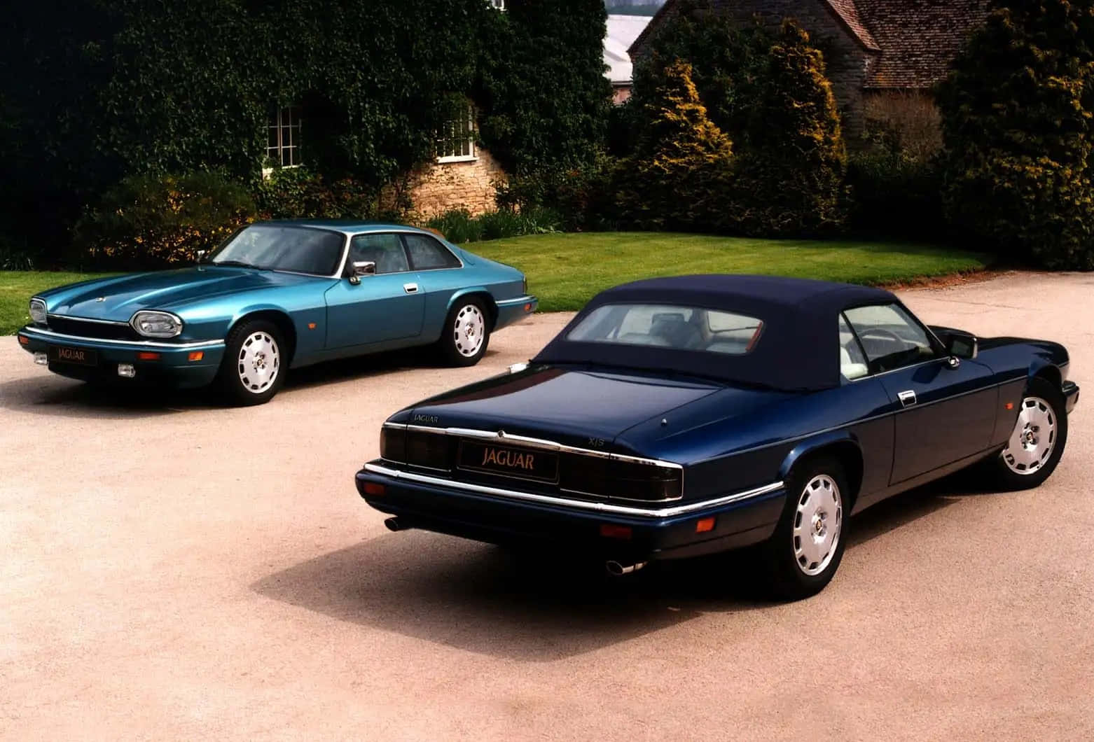 Jaguar Xjs 桌布 壁紙 — 兩輛經典積架汽車，一輛青綠色雙門轎跑車和一輛深藍色開篷車，停泊在石屋前的碎石車道上，周圍綠意盎然。