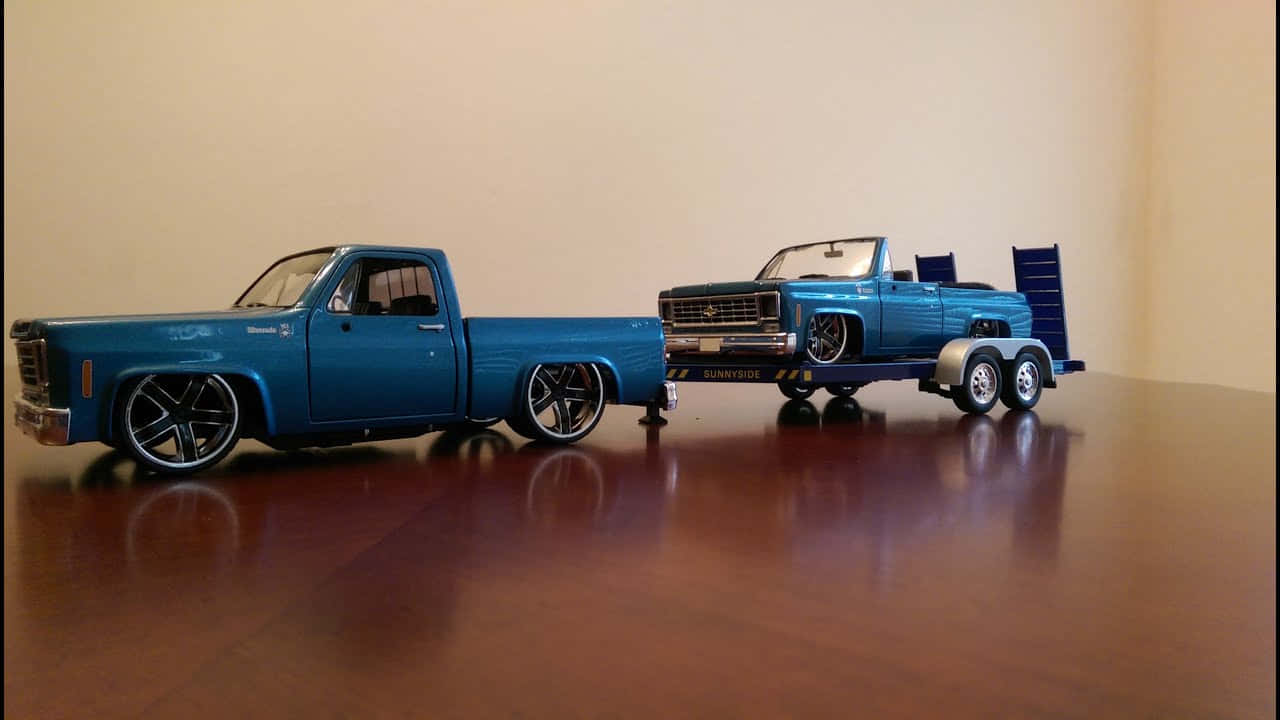 Car Model 桌布 壁紙 — 兩輛藍色定制模型卡車，一輛拖著另一輛，放在一張拋光木桌上，背景是純米色牆壁。