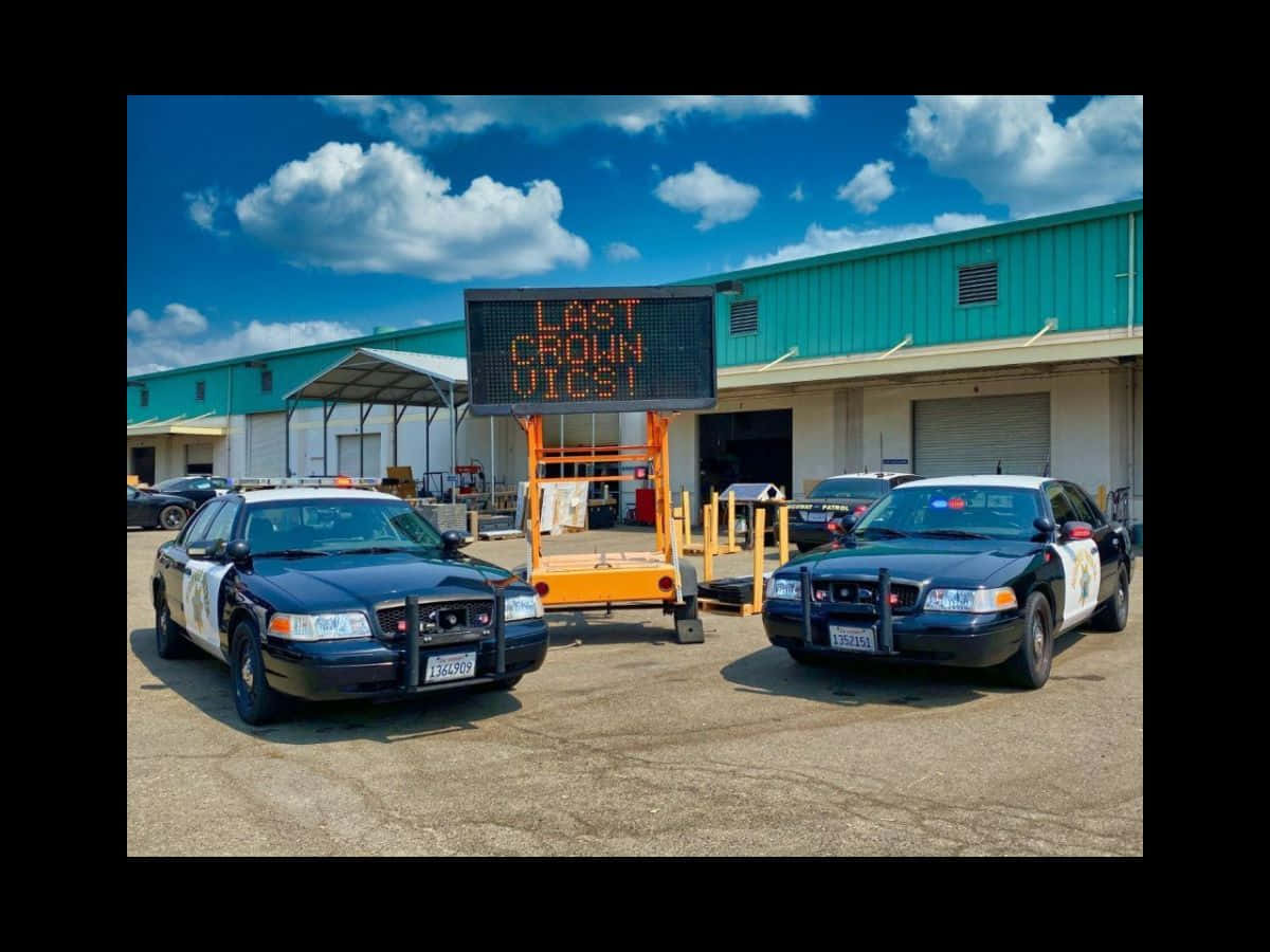 Ford Crown Victoria 桌布 壁紙 — 兩輛警車停泊在青綠色倉庫前，上方是明亮的藍天和蓬鬆的白雲。