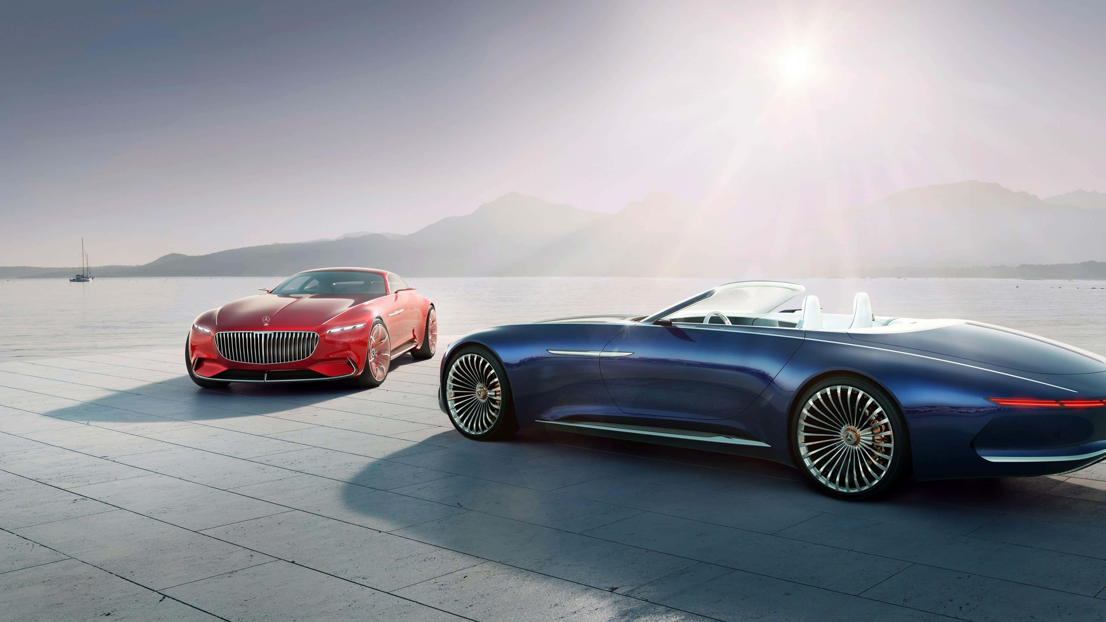Maybach 桌布 壁紙 — 兩輛豪華概念車，一輛紅色轎跑車和一輛藍色敞篷車，在明亮的陽光下閃耀，位於海濱廣場，遠處有山巒。
