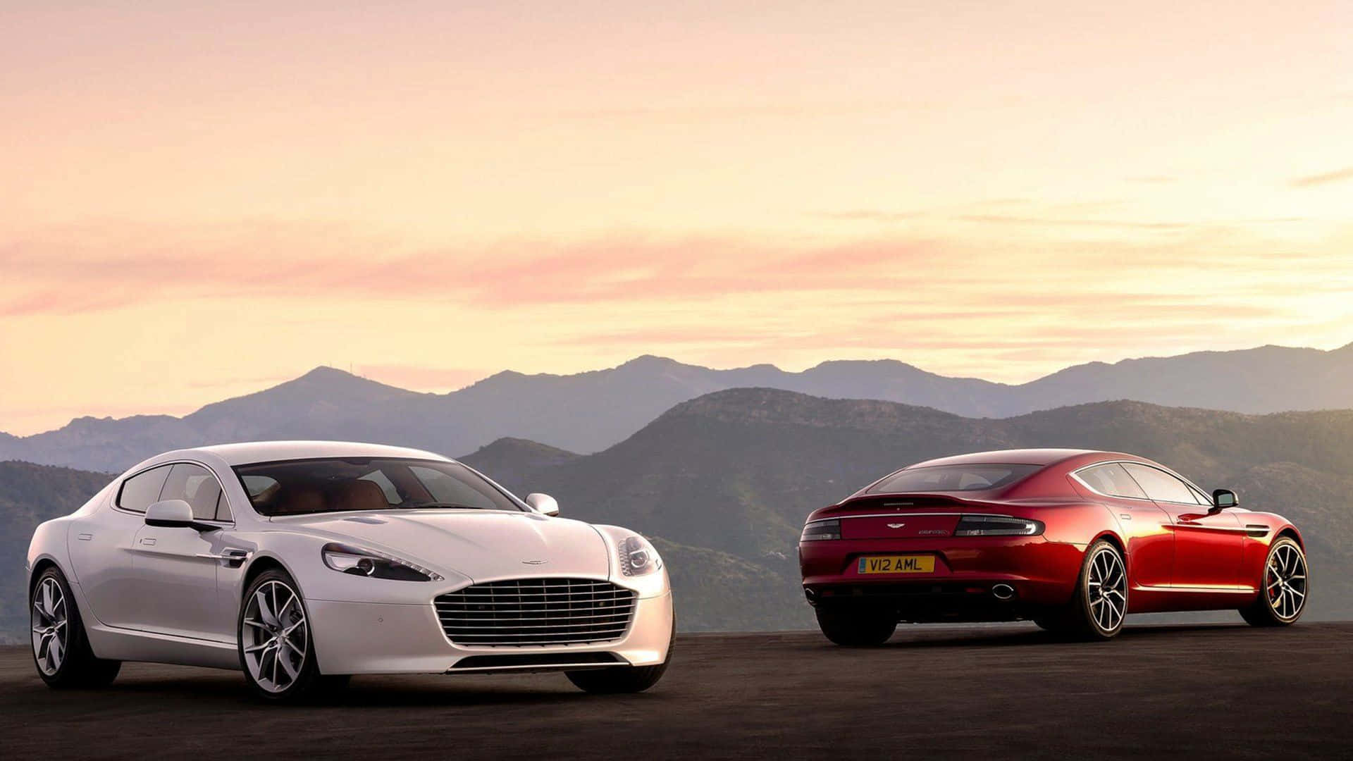 Aston Martin Rapide S 桌布 壁紙 — 兩輛豪華跑車白色和紅色停在山頂觀景點，夕陽下天空呈現柔和的粉紅色和橙色。
