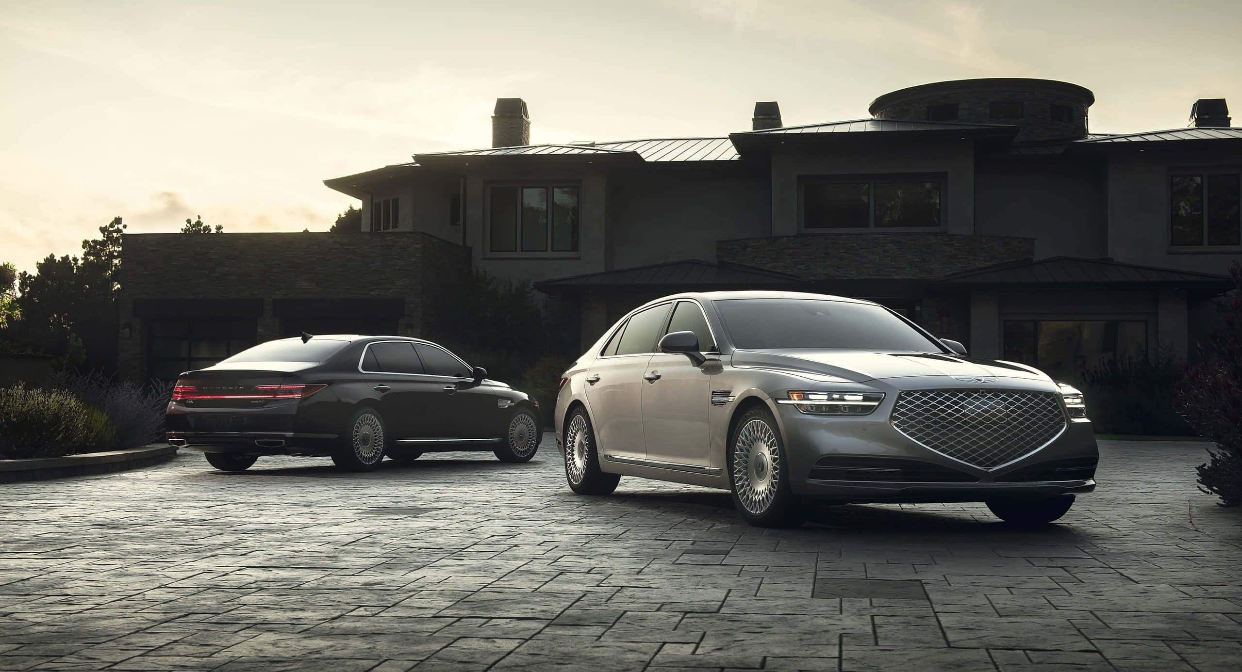 Genesis G90 桌布 壁紙 — 兩輛豪華轎車停泊在現代房屋前的石頭車道上，正值黃金時刻。