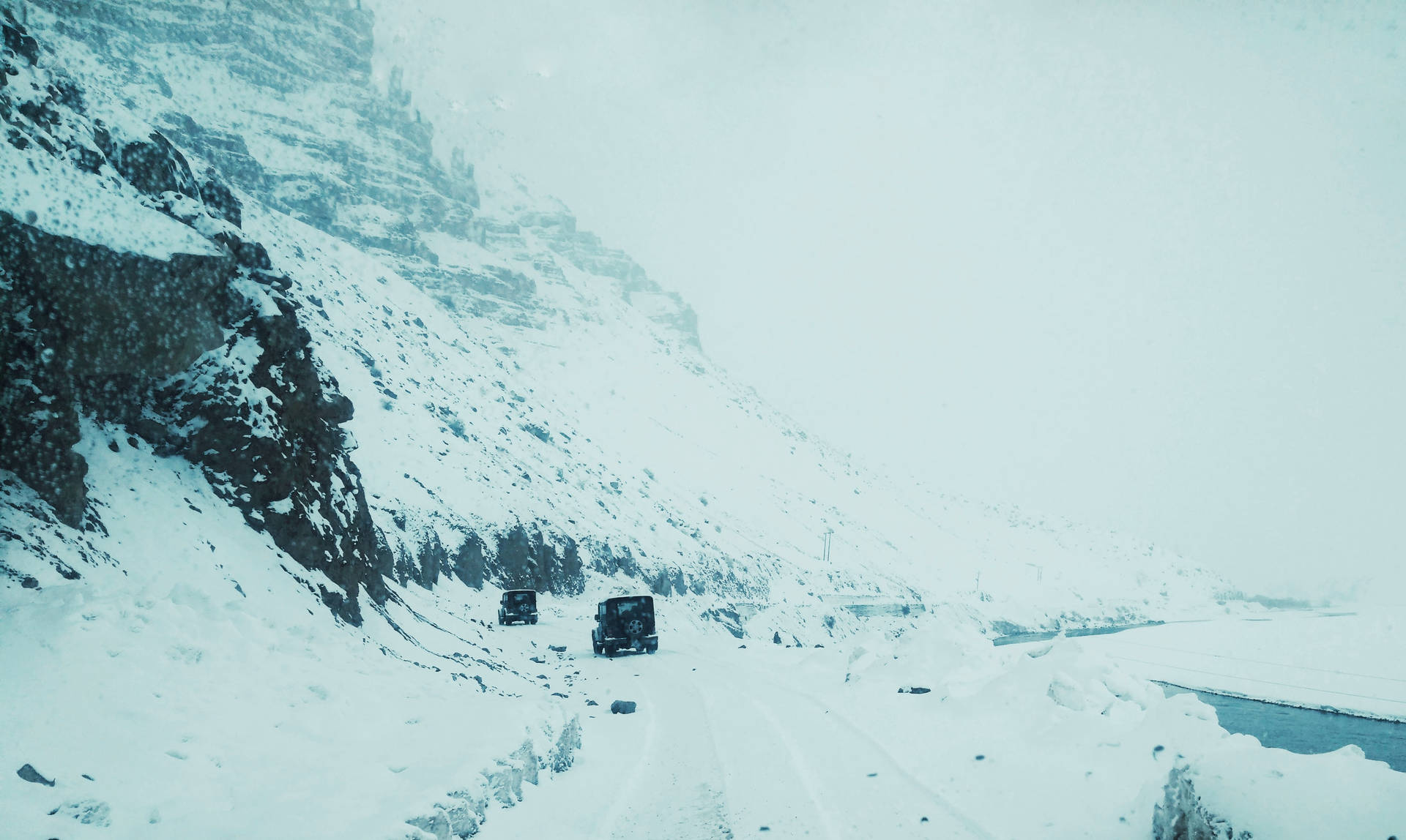  壁紙 — 兩輛貨車在暴風雪中行駛於積雪的山路上，一側是陡峭的岩石懸崖，下方是一條結冰的河流。