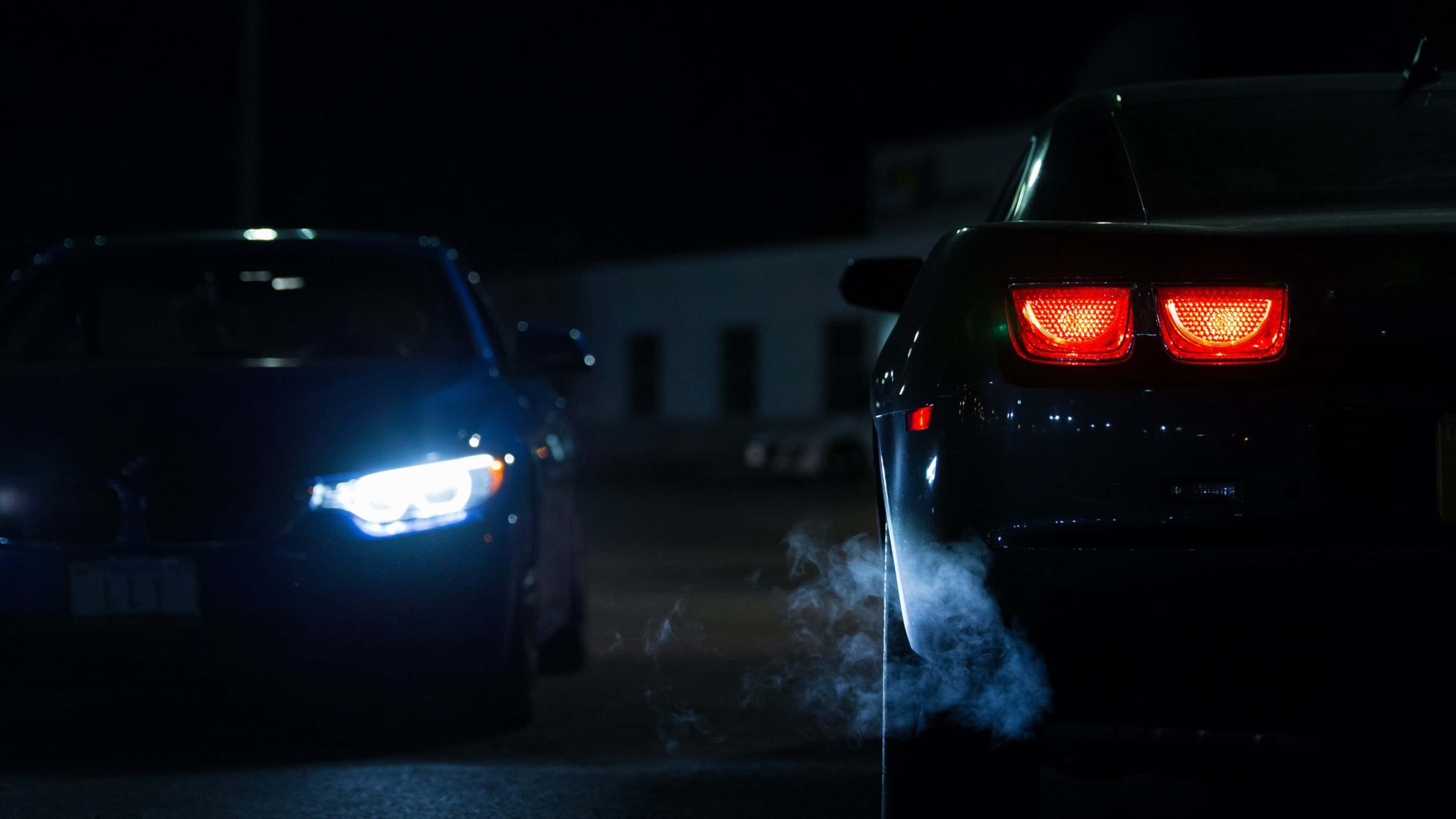 4K Car 桌布 壁紙 — 兩輛車在夜間對峙，一輛發出藍色光芒的車頭燈，另一輛則有紅色尾燈和從排氣管冒出的煙霧。
