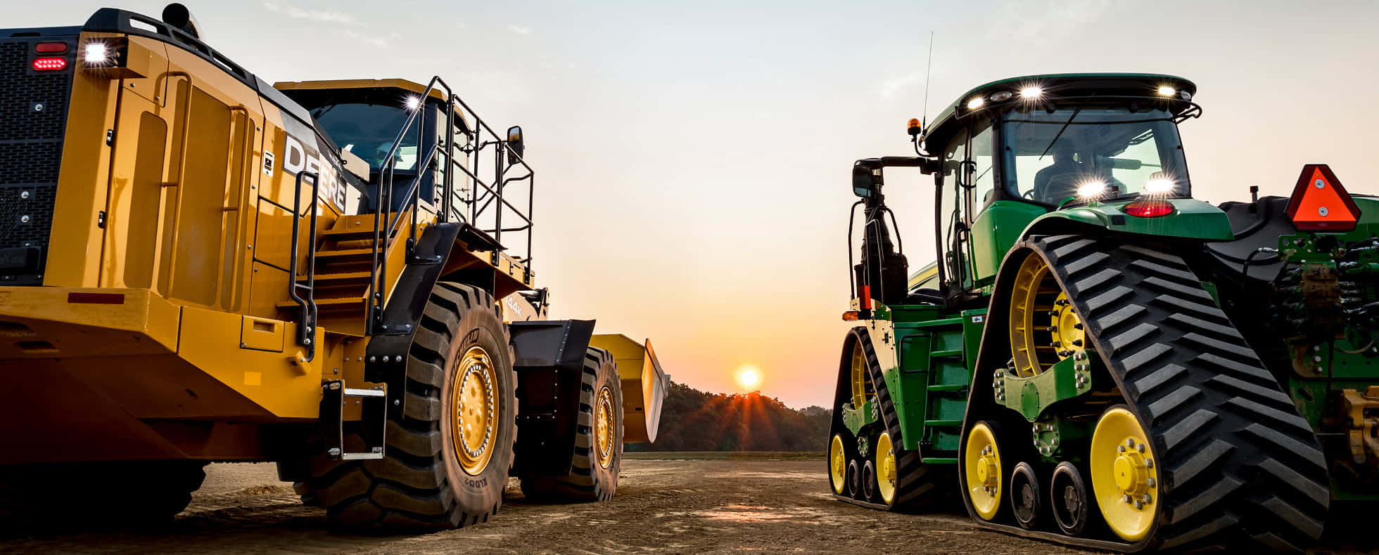 John Deere 桌布 壁紙 — 兩輛重型工程車，一輛黃色裝載機和一輛綠色拖拉機，在溫暖的日落天空下相對而立。