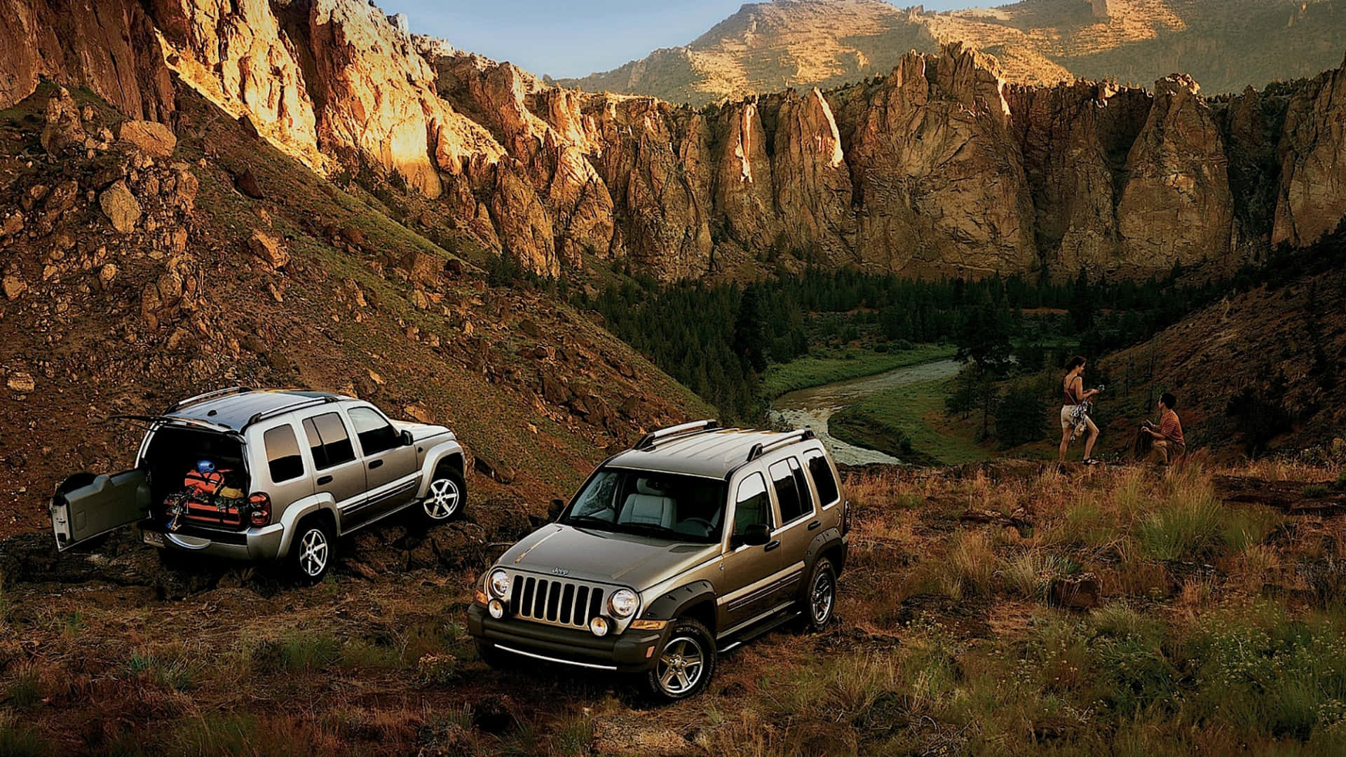 Jeep Liberty 桌布 壁紙 — 兩輛銀色Jeep停泊在崎嶇的峽谷小徑上，遠足者正在探索下方風景如畫的山谷。