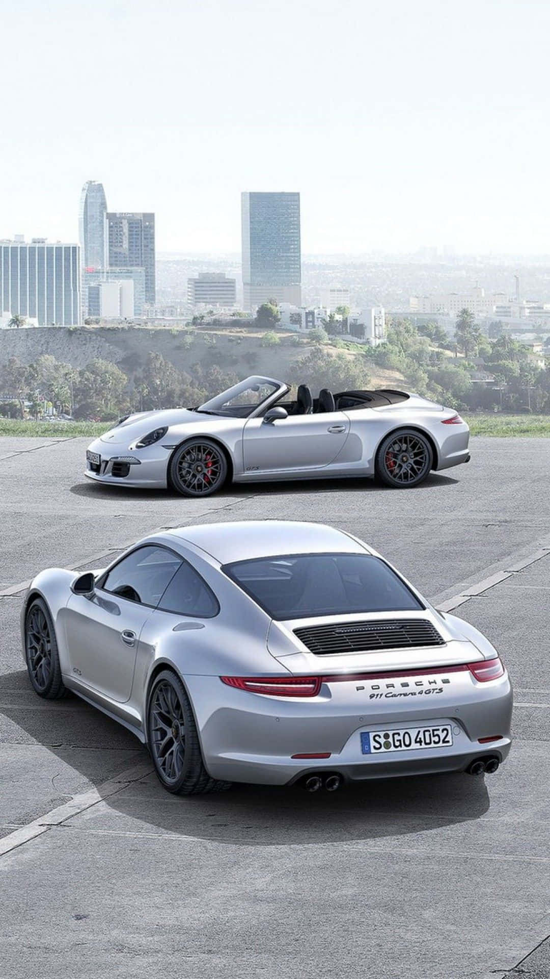 Porsche iPhone 桌布 壁紙 — 兩輛銀色保時捷911 Carrera GTS跑車停泊在混凝土觀景台上，背景是朦朧的城市天際線。
