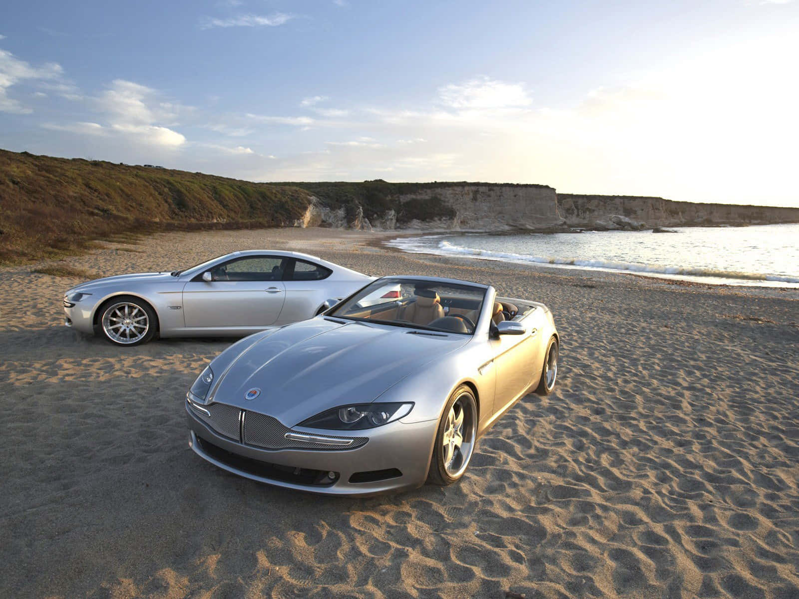 Fisker 桌布 壁紙 — 兩輛銀色豪華開篷車停泊在陽光明媚的沙灘上，背景有懸崖和海洋波浪。
