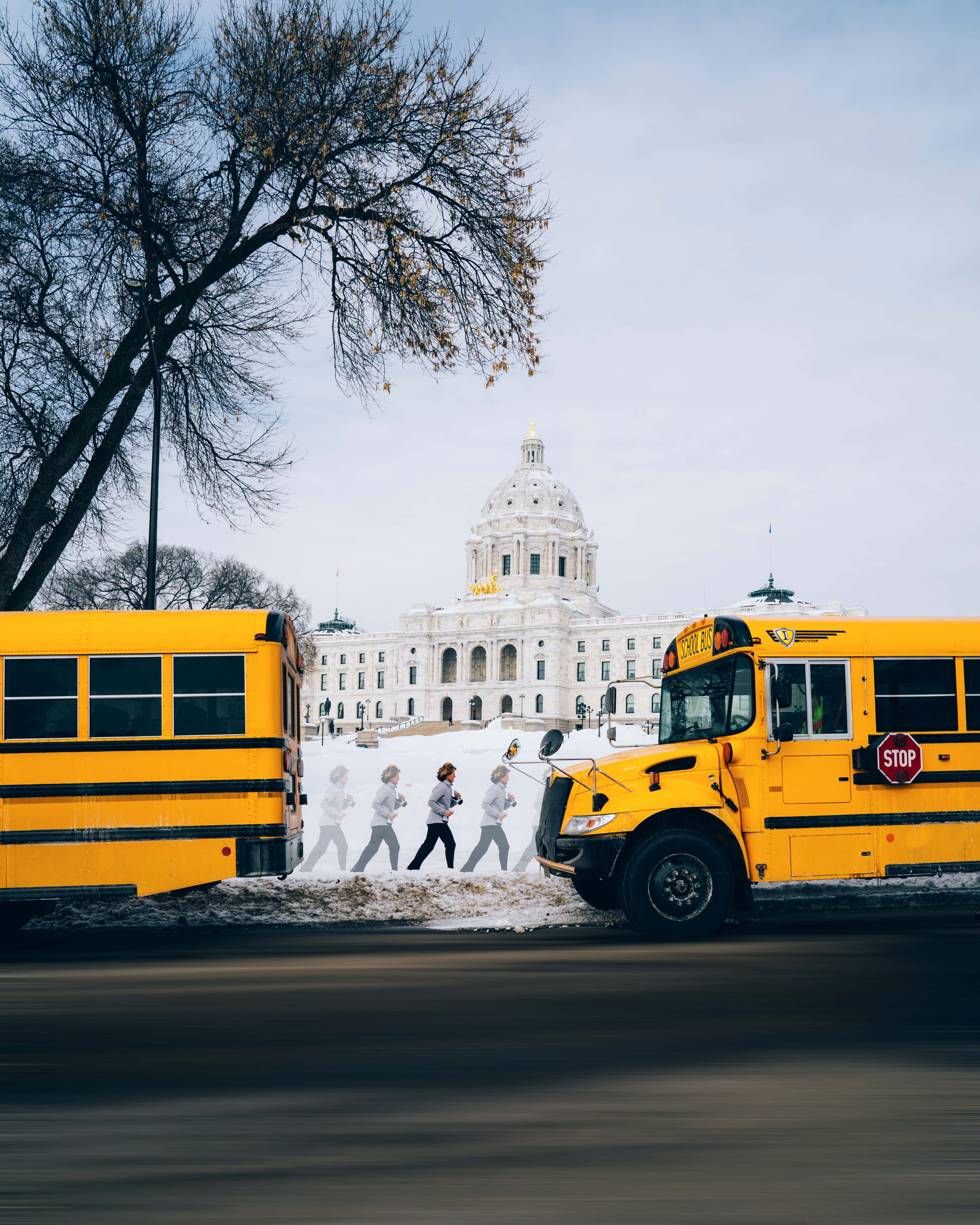 Yellow School Bus 桌布 壁紙 — 兩輛黃色校車框住一條積雪街道，背景是宏偉的白色國會大廈和光禿禿的樹木，天空多雲。