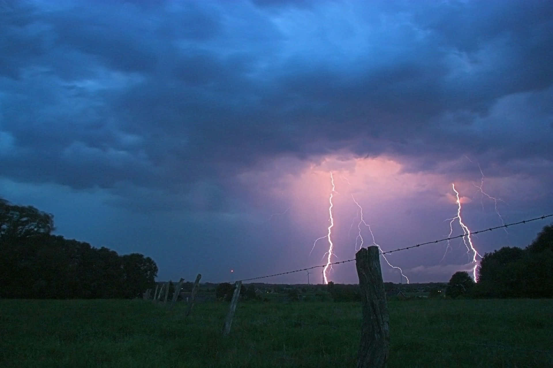 Thunderstorm 桌布 壁紙 — 兩道鋸齒狀閃電照亮了暴風雨黃昏的天空，下方是帶有帶刺鐵絲網圍欄的草地。