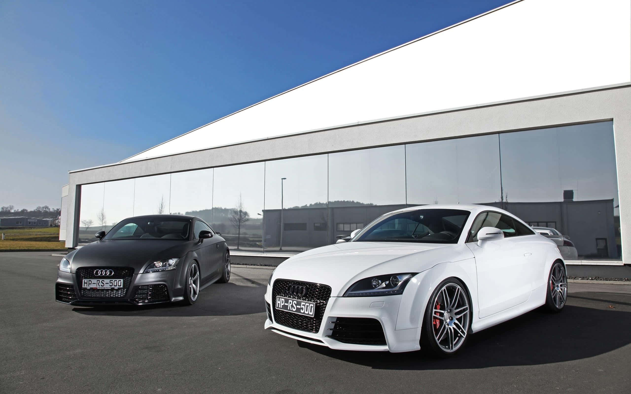 Audi Tt Rs 桌布 壁紙 — 兩部高性能奧迪 TT RS 500 雙門跑車，一部啞光黑色，一部亮光白色，停泊在現代玻璃建築前，天空清澈湛藍。