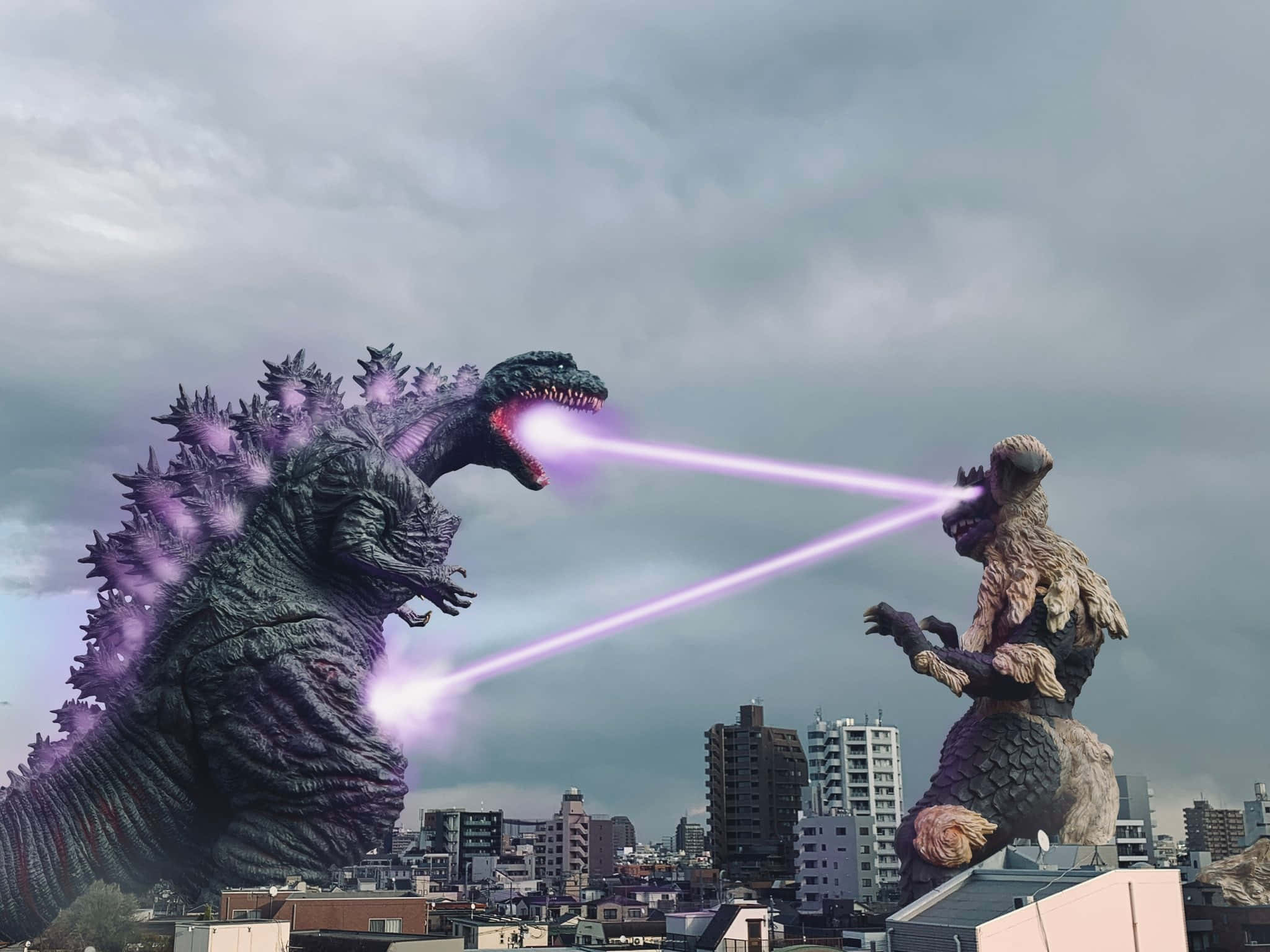 Godzilla Vs King Caesar 桌布 壁紙 — 兩隻巨型怪獸哥斯拉和王者基多拉在城市天際線上戰鬥，向多雲的天空發射紫色原子吐息。