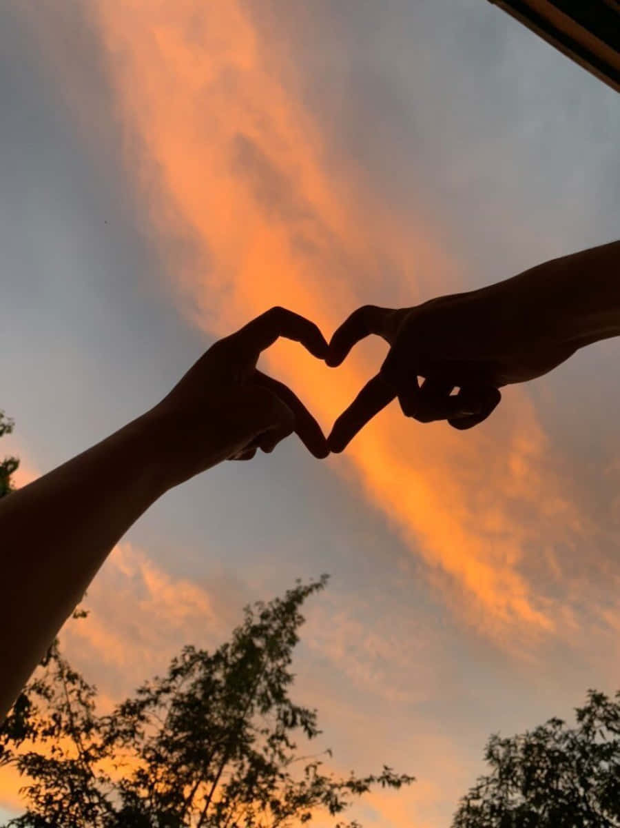 Heart Silhouette 桌布 壁紙 — 兩隻手在充滿活力的橙色日落天空下形成心形，下方有樹木的剪影。