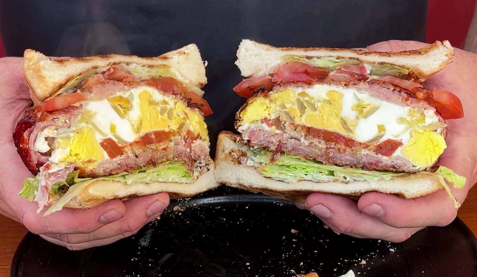 bacon 桌布 壁紙 — 兩隻手自豪地展示一個新鮮切開的煙肉蛋芝士三文治，露出融化芝士、火腿、番茄和生菜的層次。